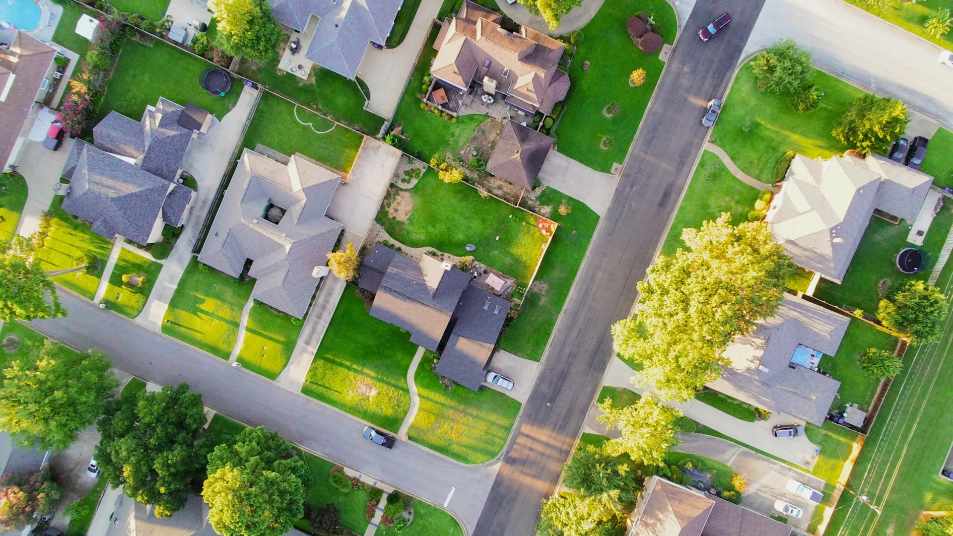 Aerial view of a suburban neighborhood with houses, green lawns, trees, and roads on a sunny day.