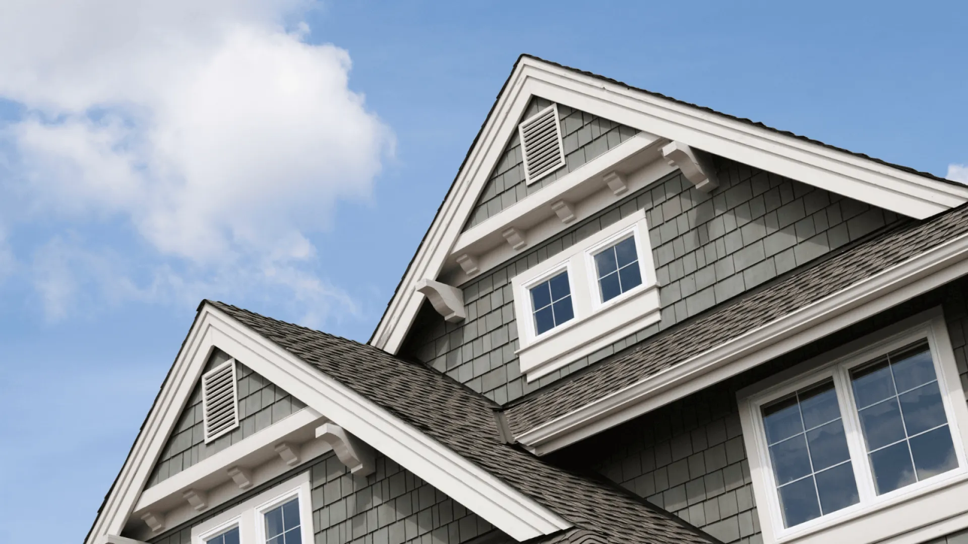 Close-up of grey shingle roof with white trimmed gable windows against a clear blue sky.