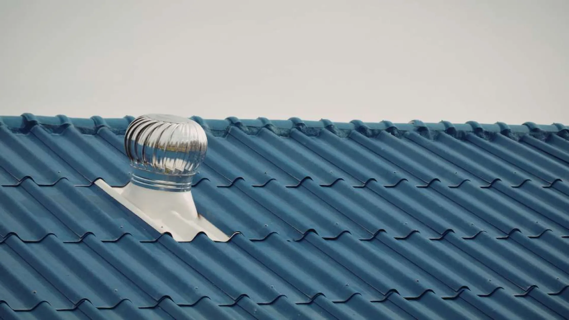 Silver metal turbine roof vent on blue corrugated metal roofing under clear sky.
