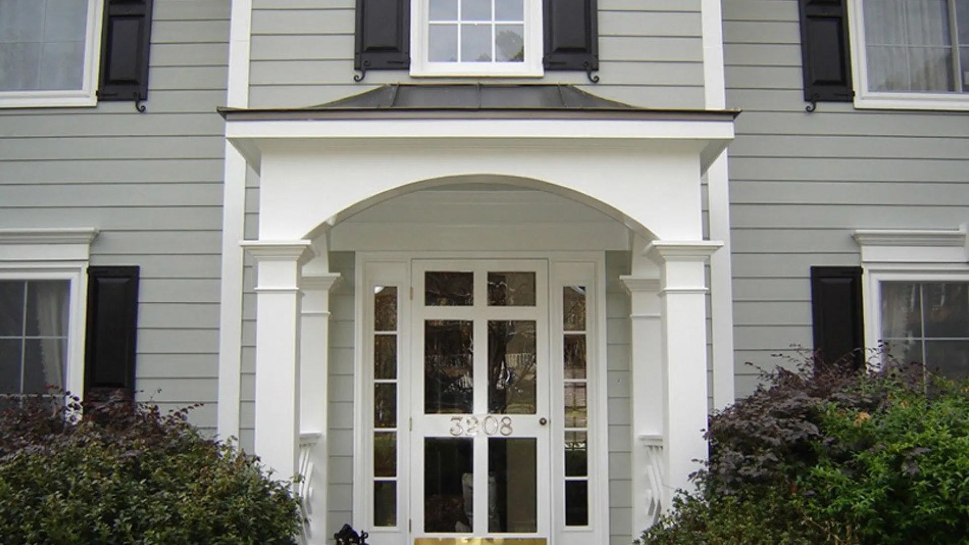 Elegant home entrance with a white portico, black shutters, and lush greenery framing the doorway.