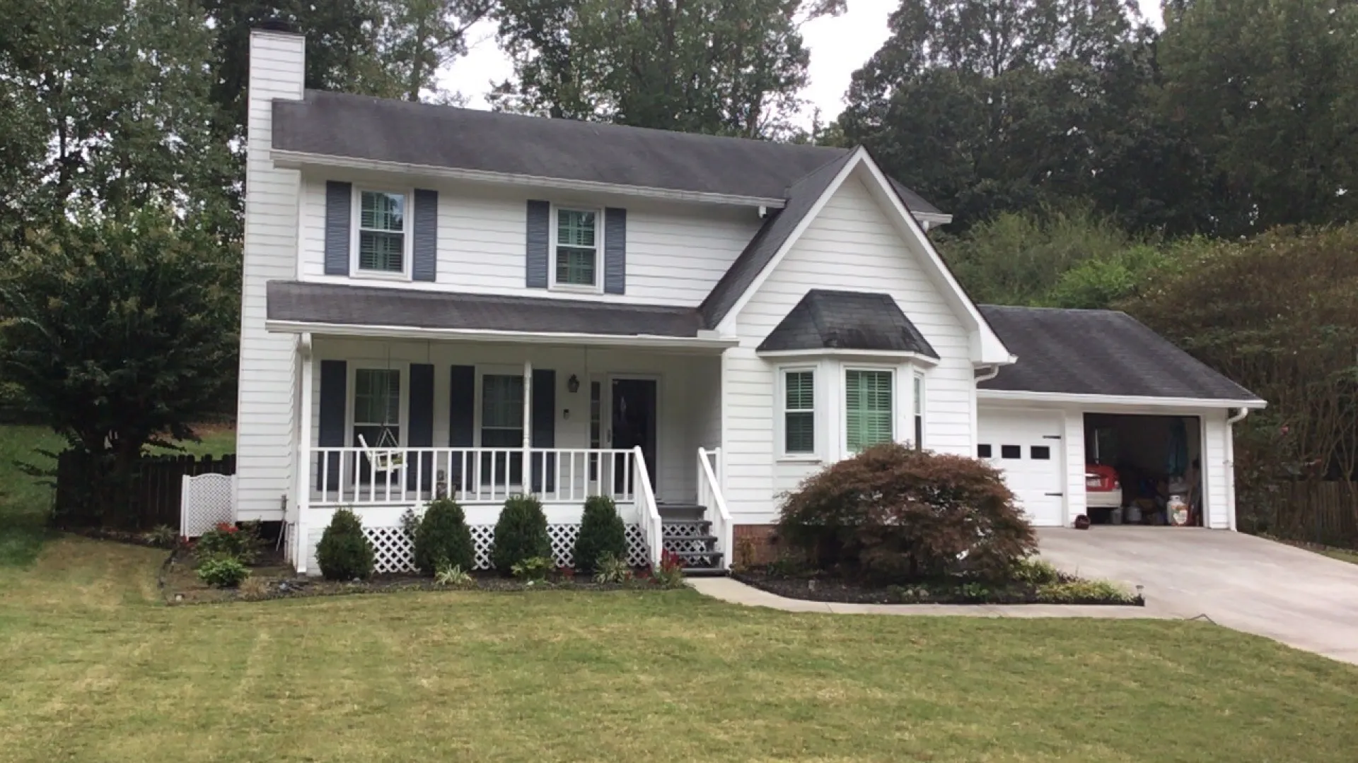 Charming two-story white house with a porch, green lawn, and garage surrounded by trees.