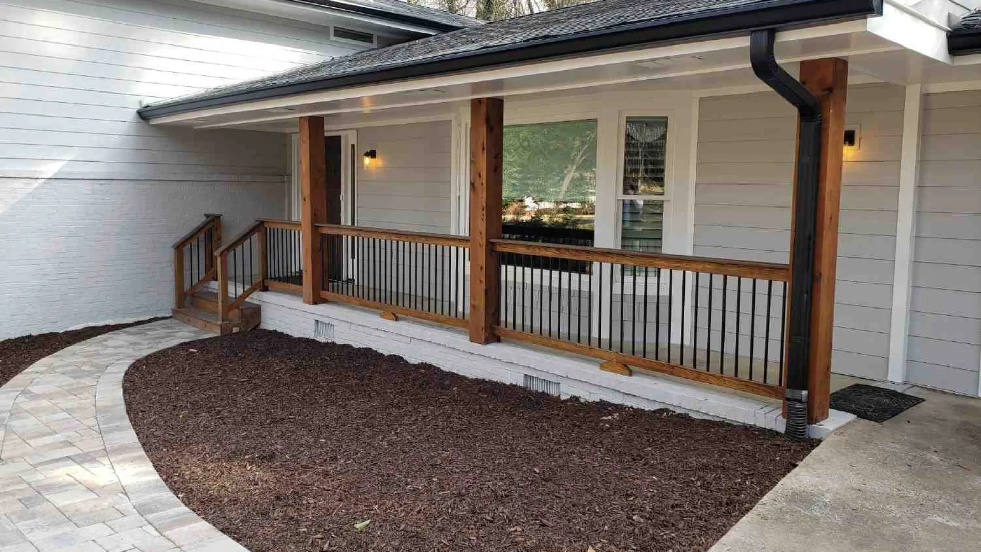 Modern front porch with wooden posts, black metal railings, and stone paver pathway around a mulch bed.