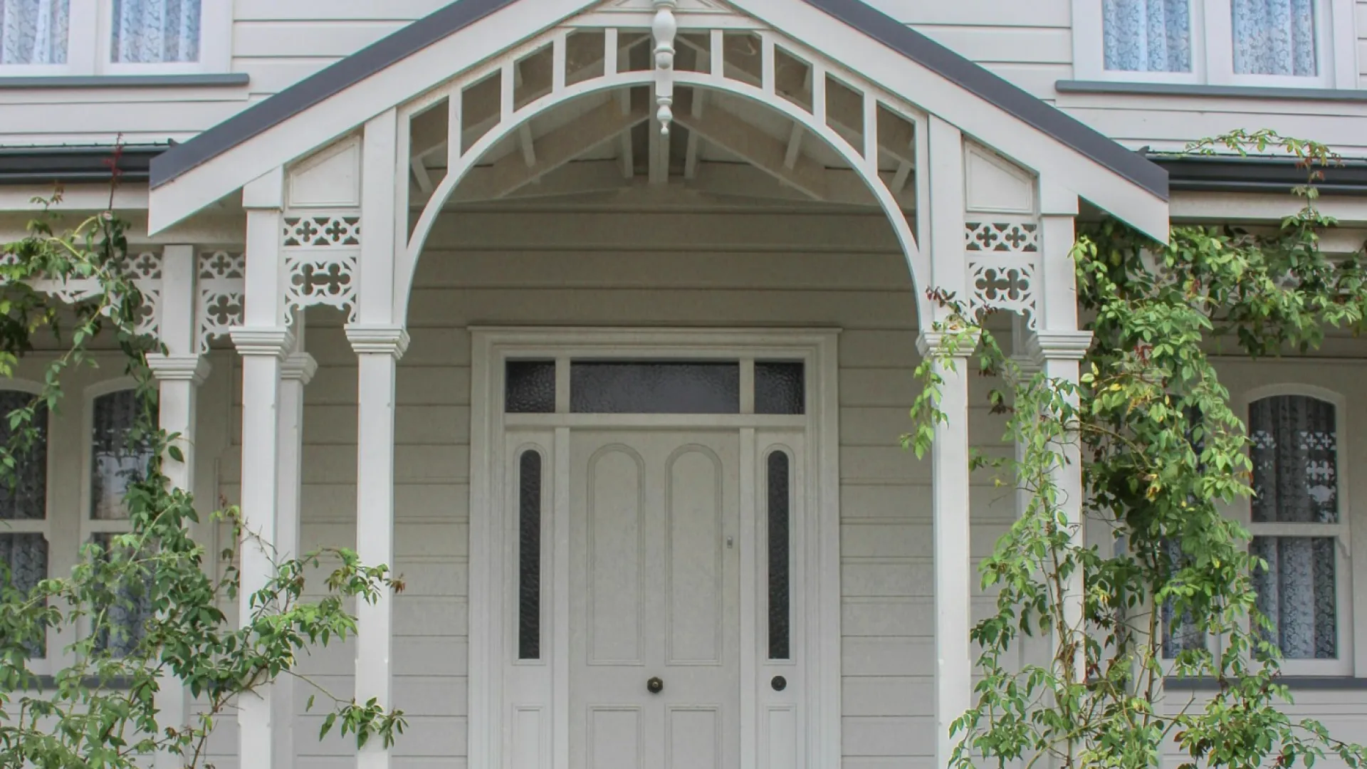 White wooden Victorian house entrance with detailed porch, stairs, and surrounding greenery and flowers.