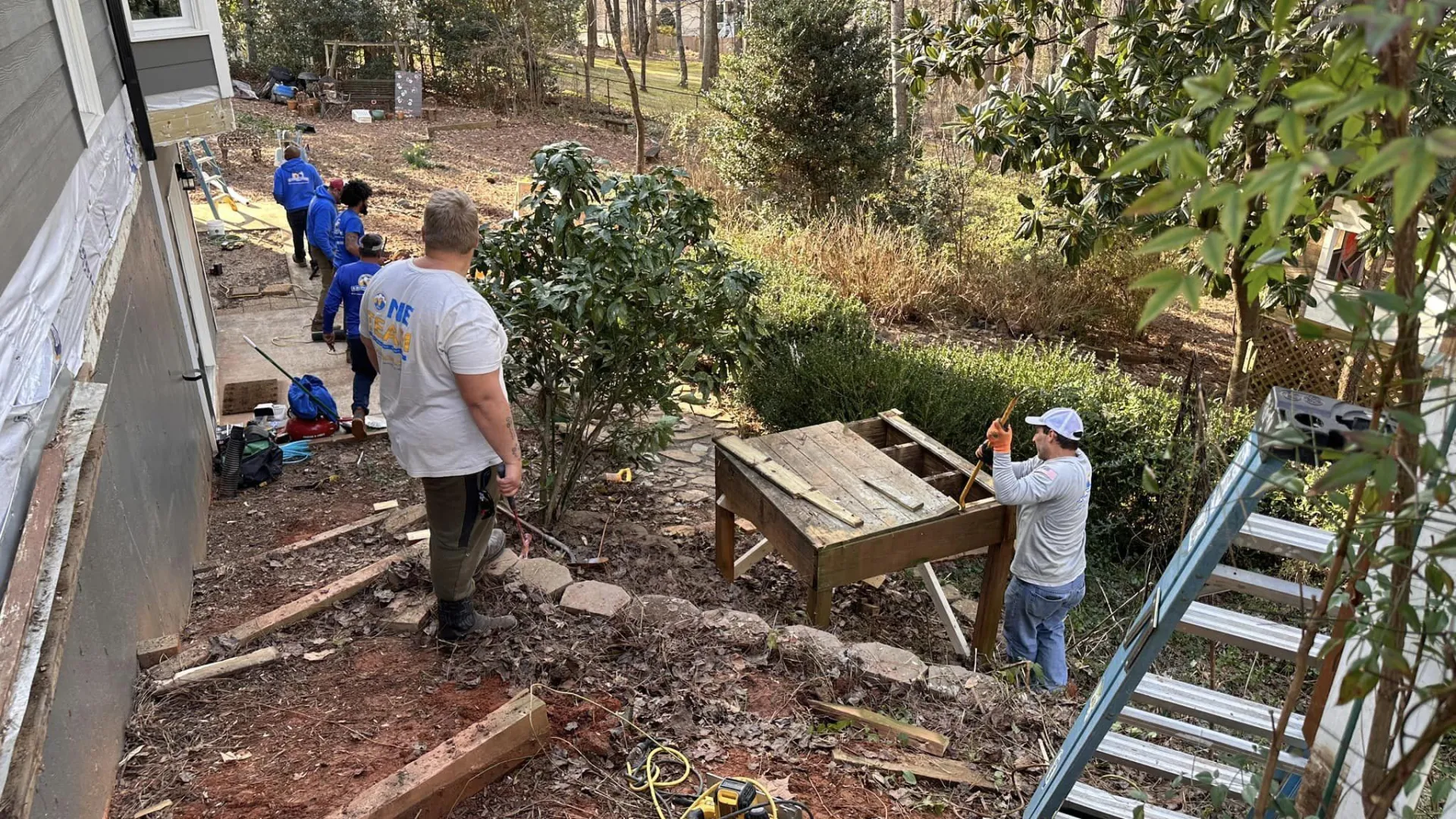 Workers building a wooden deck in a backyard surrounded by trees and garden tools during daytime.