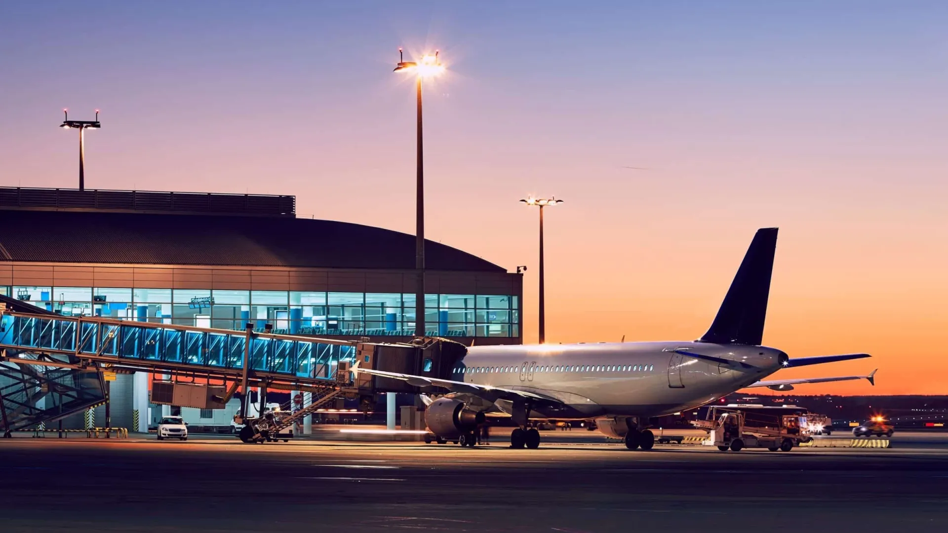 Commercial airplane parked at airport gate with jet bridge during sunset and terminal building in background