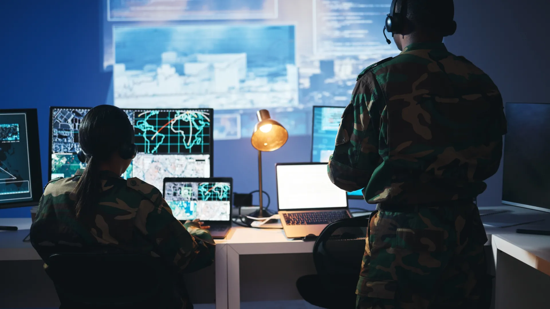 Military personnel monitoring surveillance data on multiple computer screens in a dark command center.