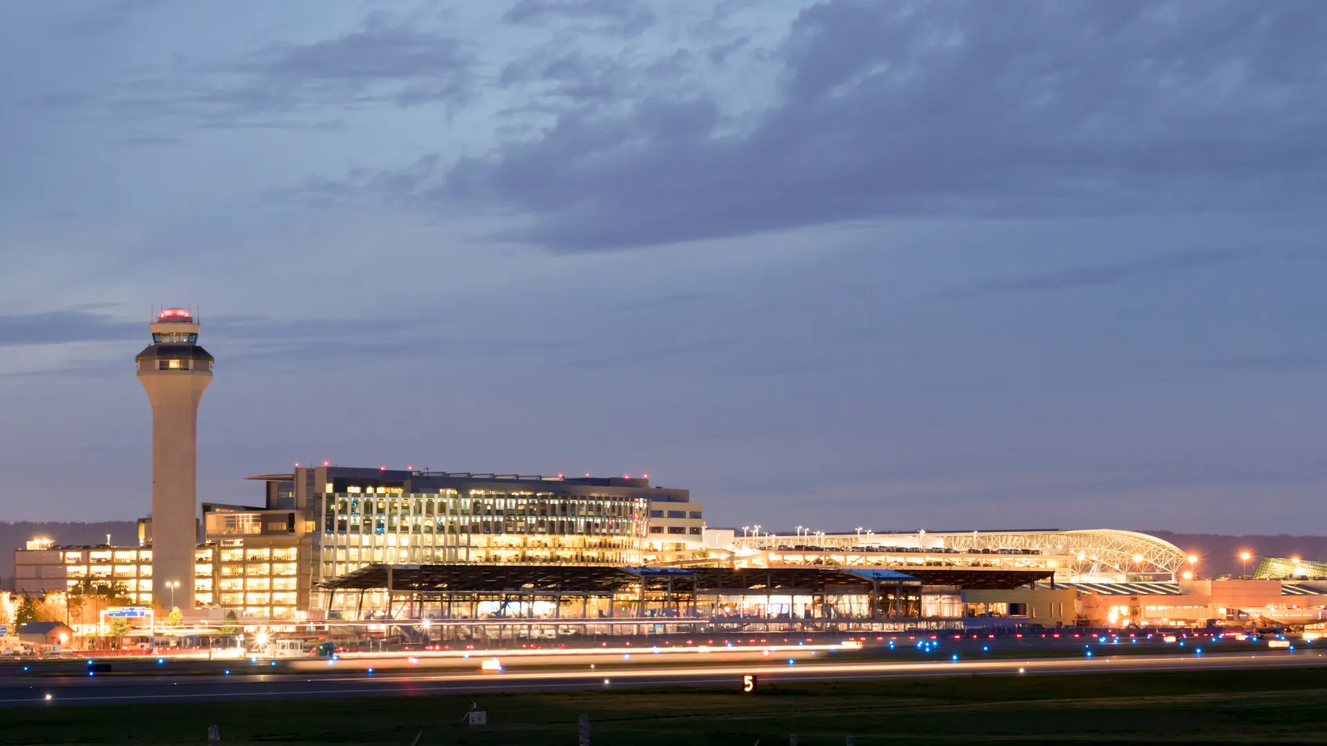 Night view of illuminated airport terminal and control tower under a cloudy sky with runway lights in foreground