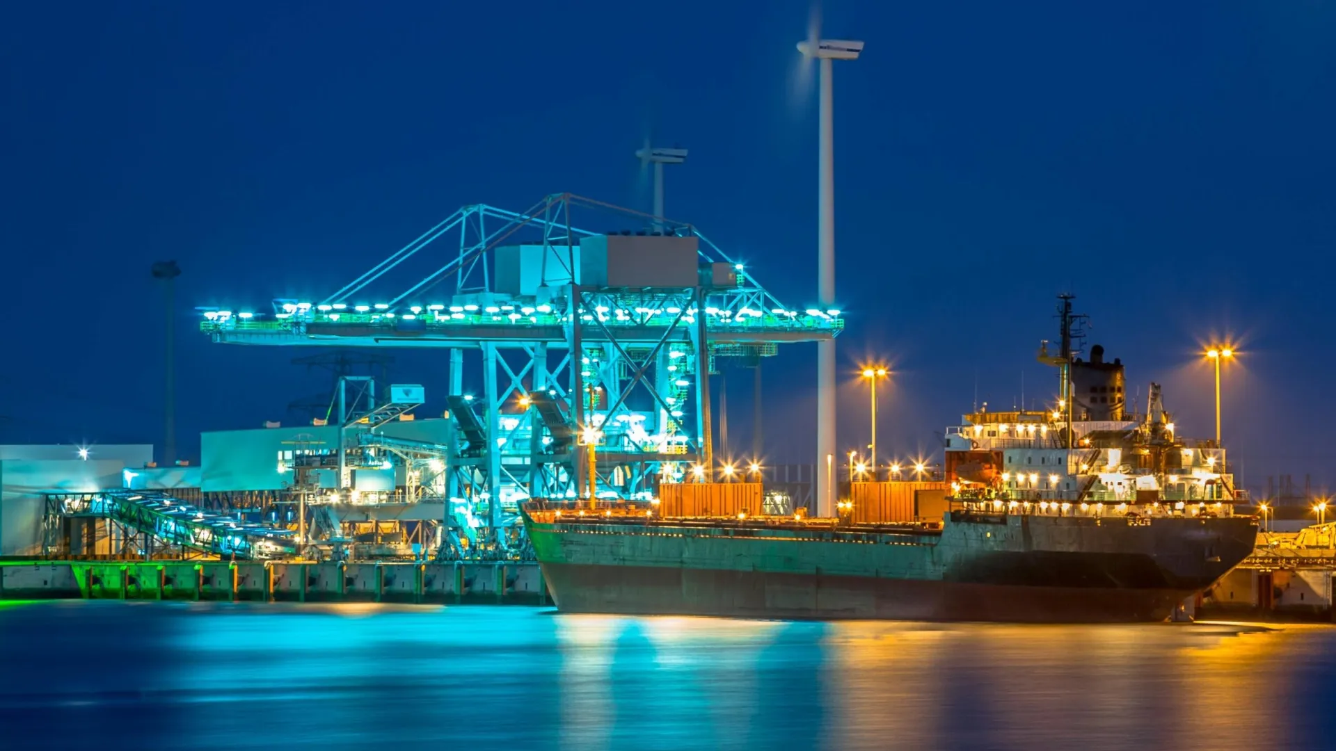 Cargo ship docked at illuminated port terminal with cranes and containers during nighttime.