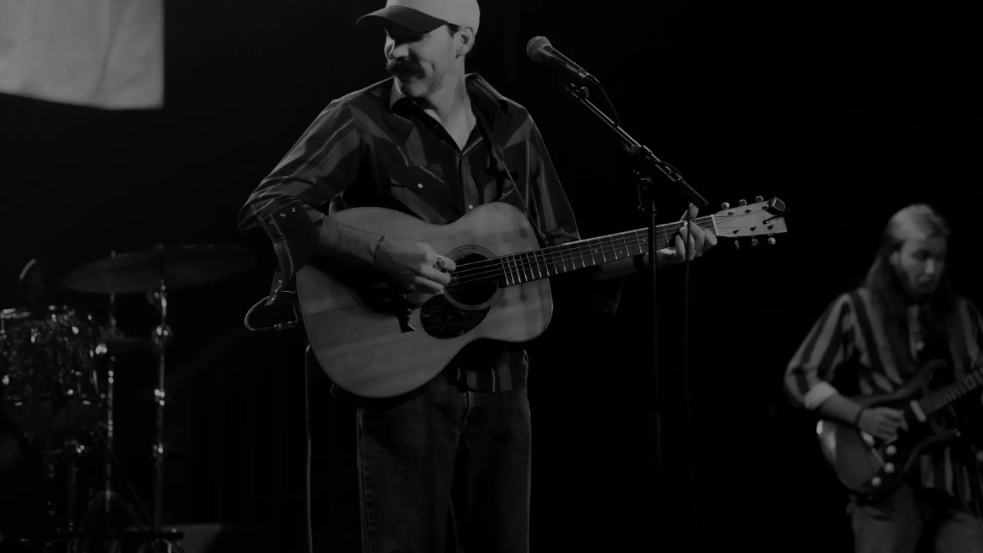 Black and white image of two musicians performing on stage with guitars and stage lights overhead.