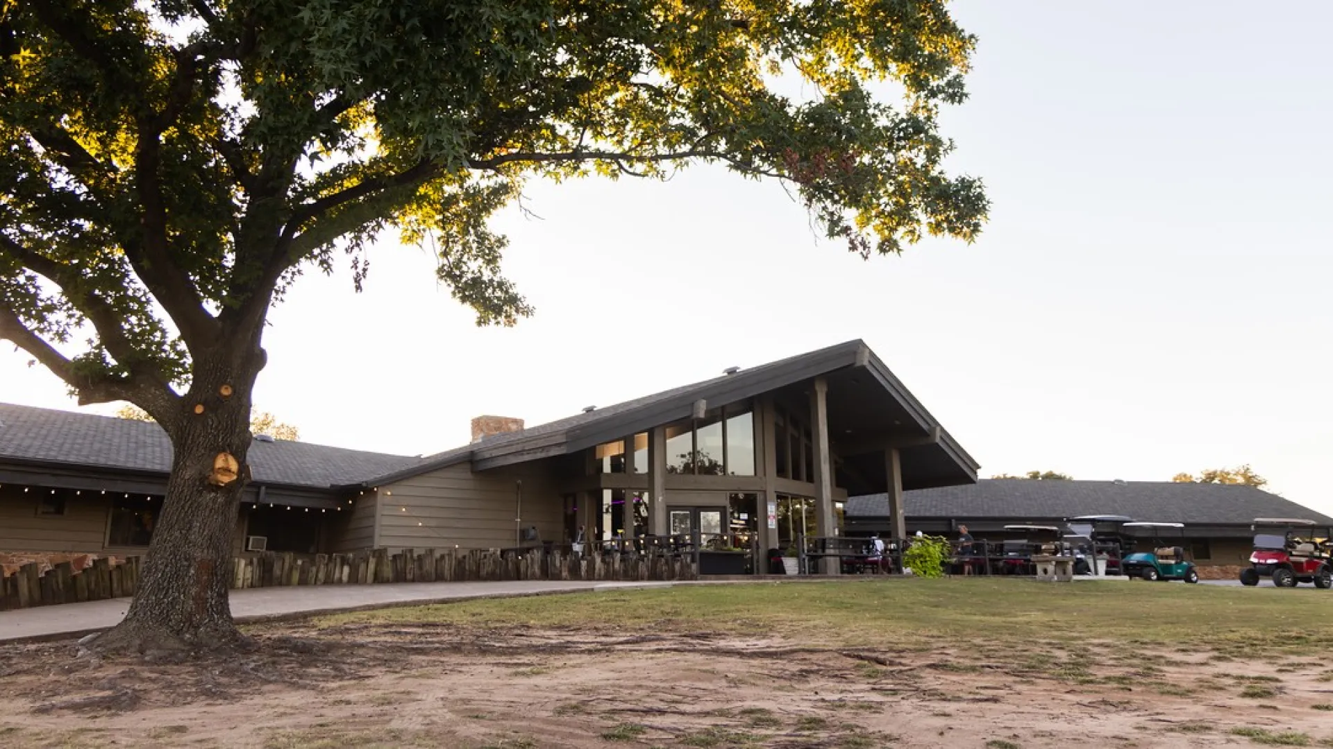Large tree next to a modern lodge-style building with a spacious lawn and golf carts outside on a clear day