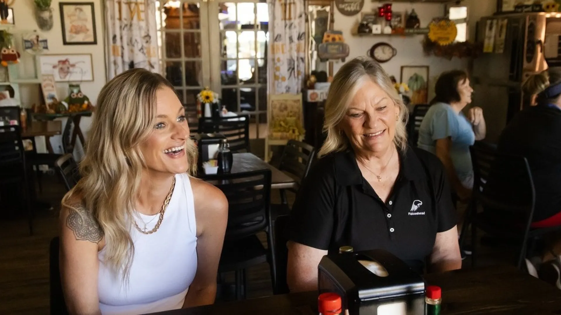 Two women smiling and sitting at a wooden table in a cozy restaurant with condiments and napkins on the table.