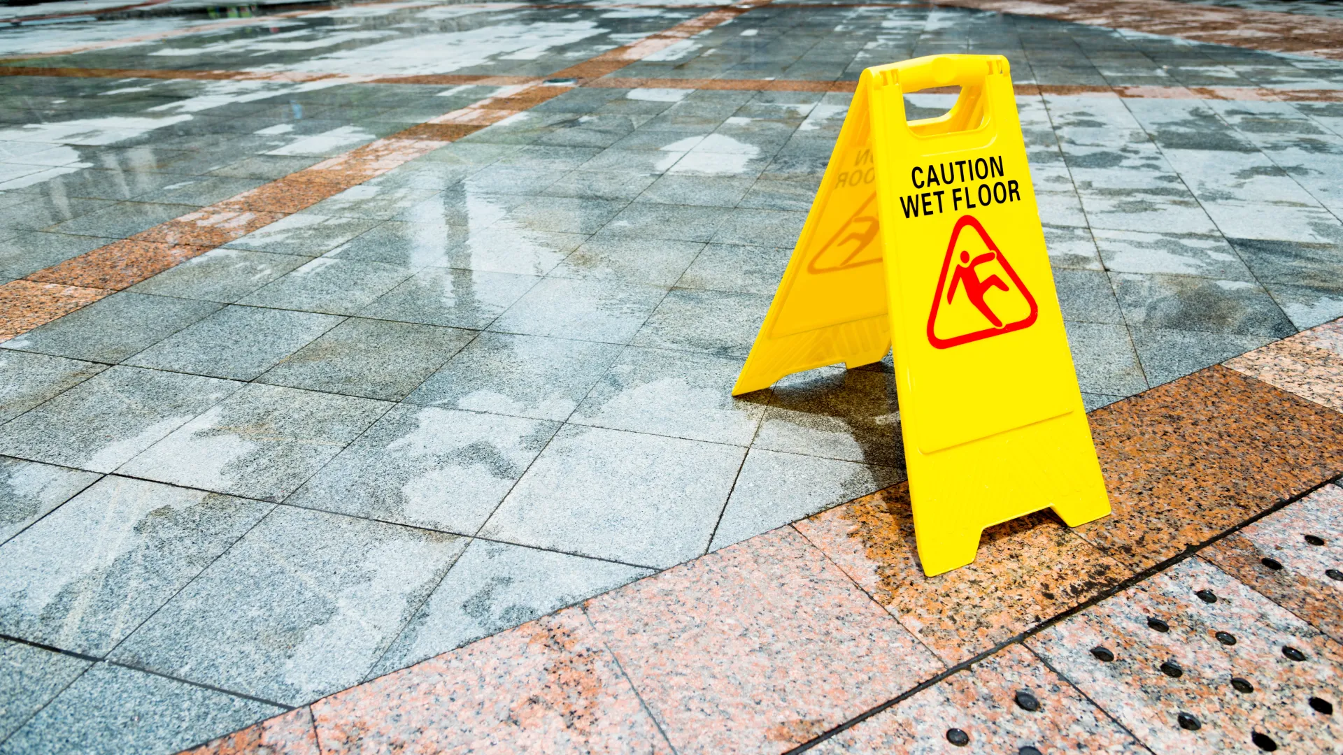 Yellow caution sign on wet tiled floor warning of slippery surface outdoors on a rainy day.