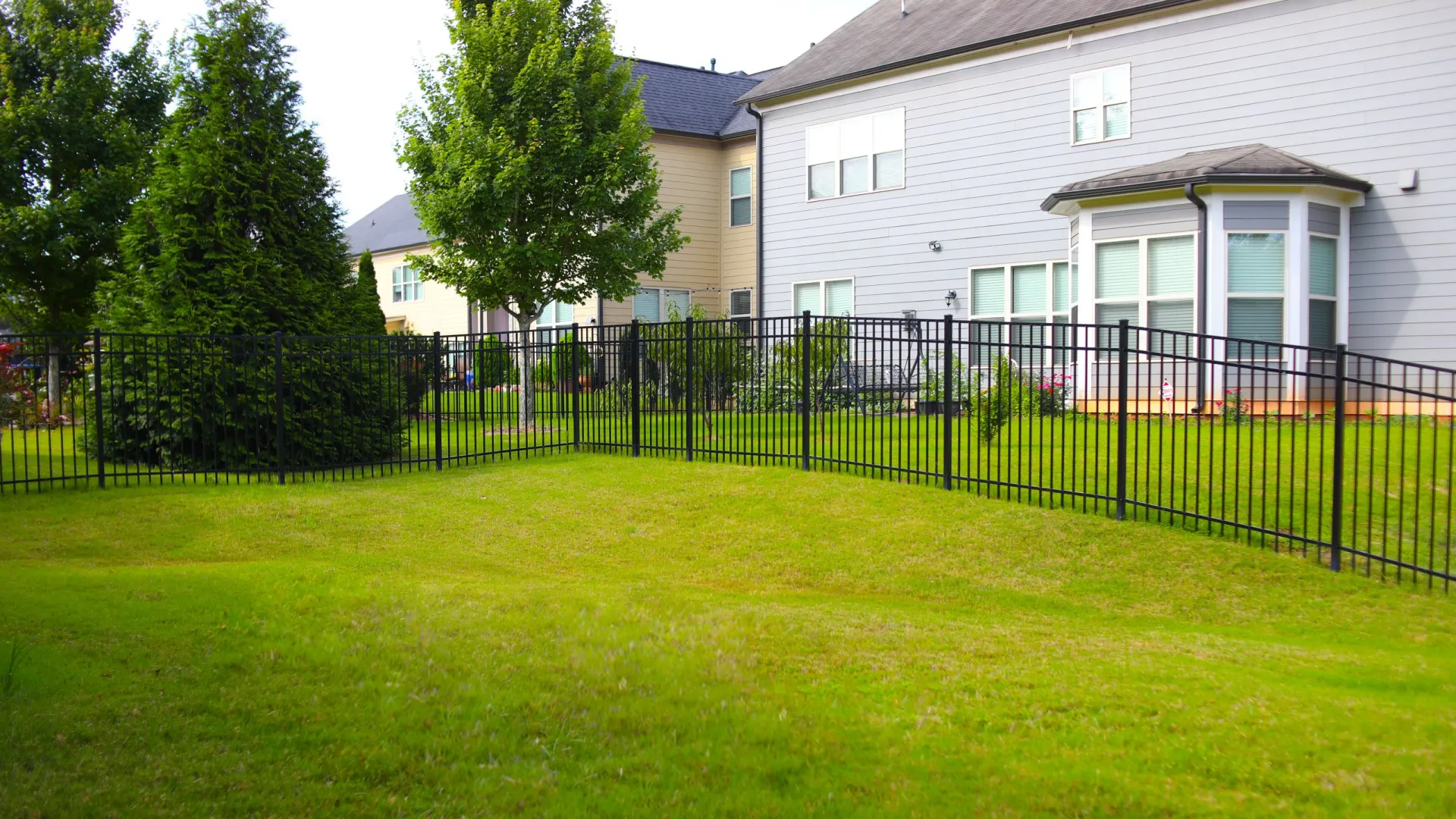 Black metal fence enclosing a green backyard with trees and suburban houses in the background
