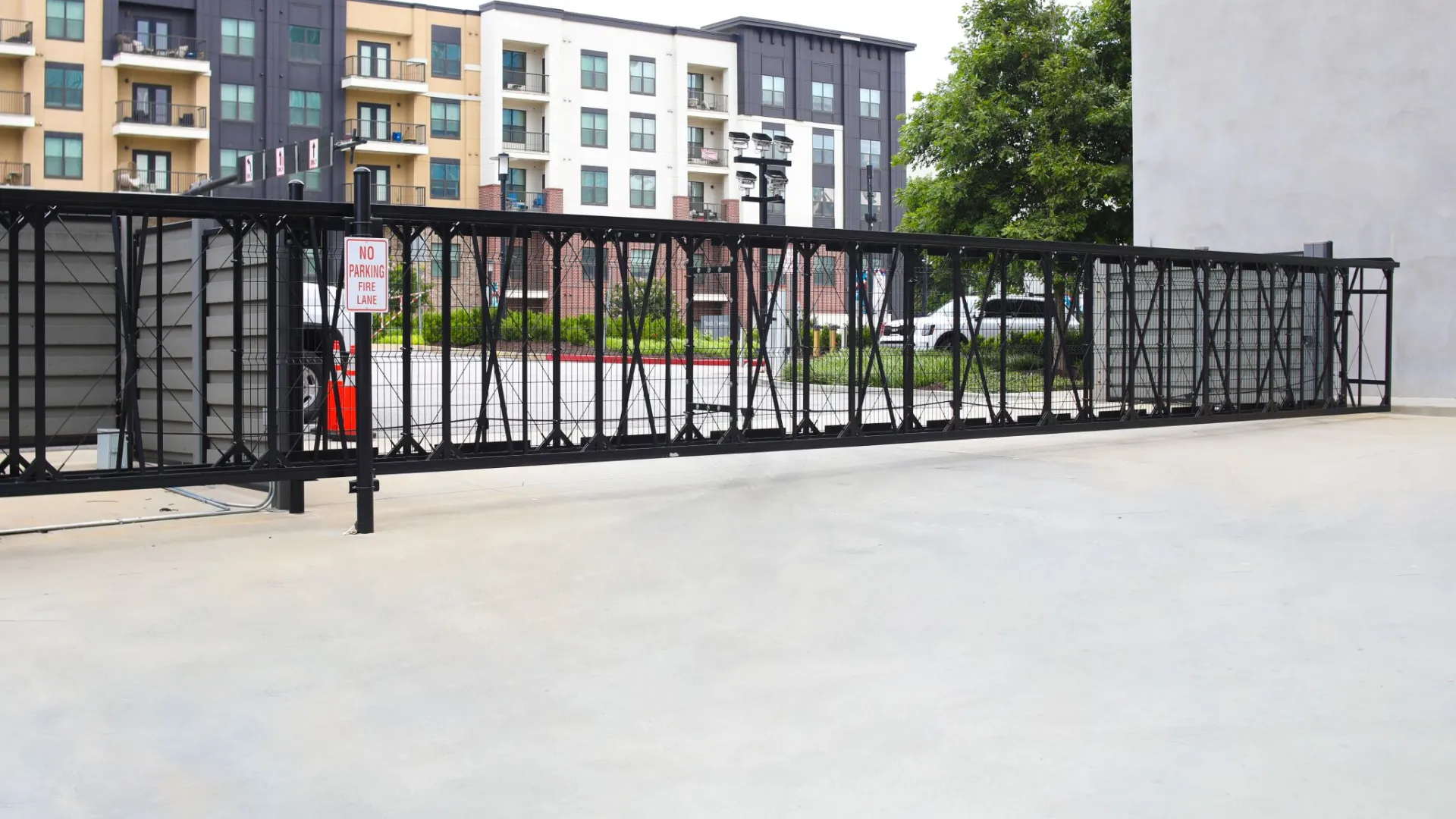 Black metal sliding security gate in front of a modern apartment building with parking lot behind it.