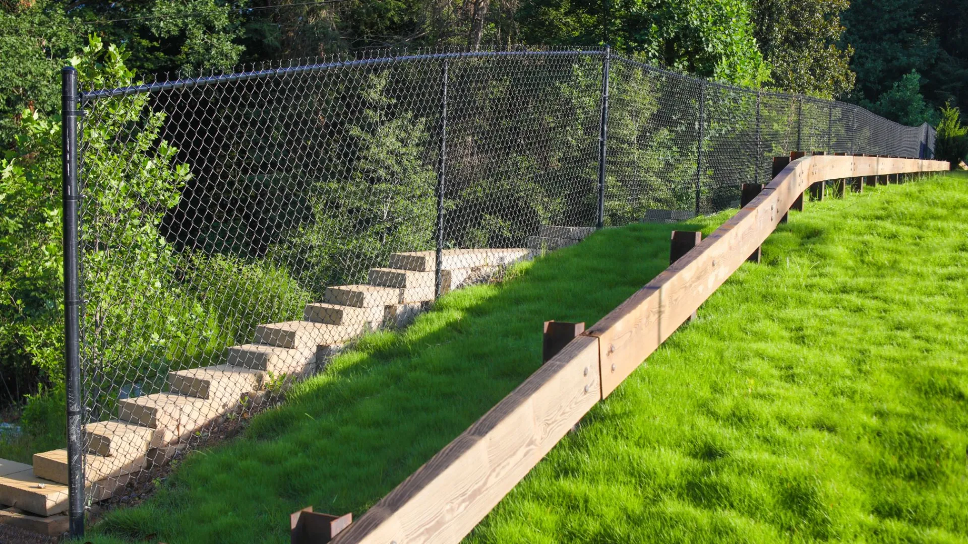 Green grassy hillside supported by wooden retaining wall with black chain-link fence and concrete steps behind it