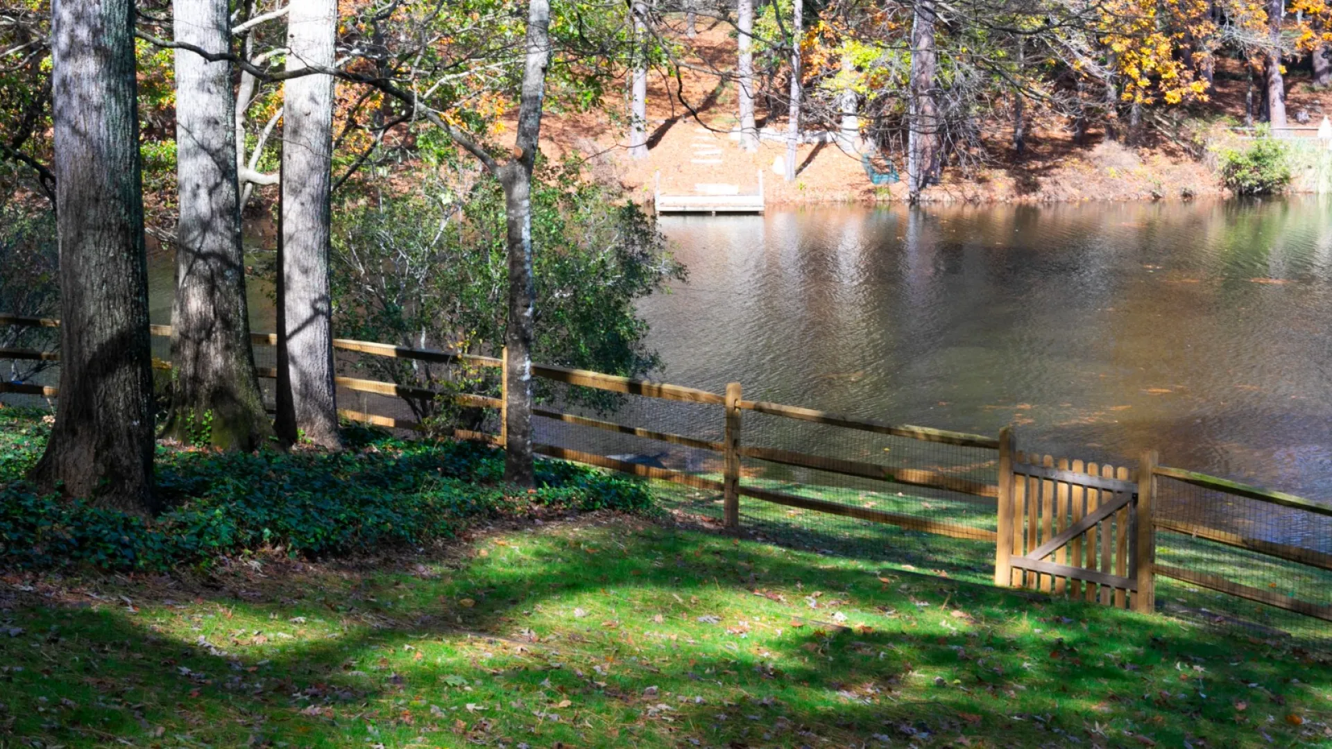 Serene lakeside view with wooden fence, autumn foliage, and reflections in the water under bright sunlight.