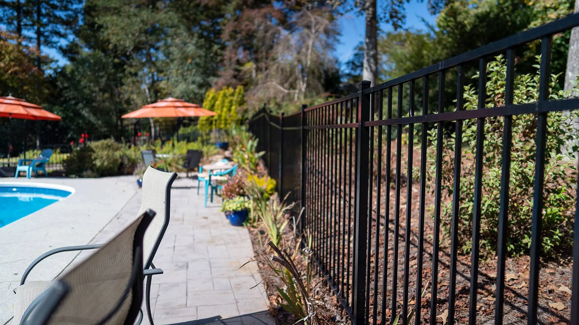 Sunny backyard pool area with a black fence, lounge chairs, and orange umbrellas under a clear blue sky.