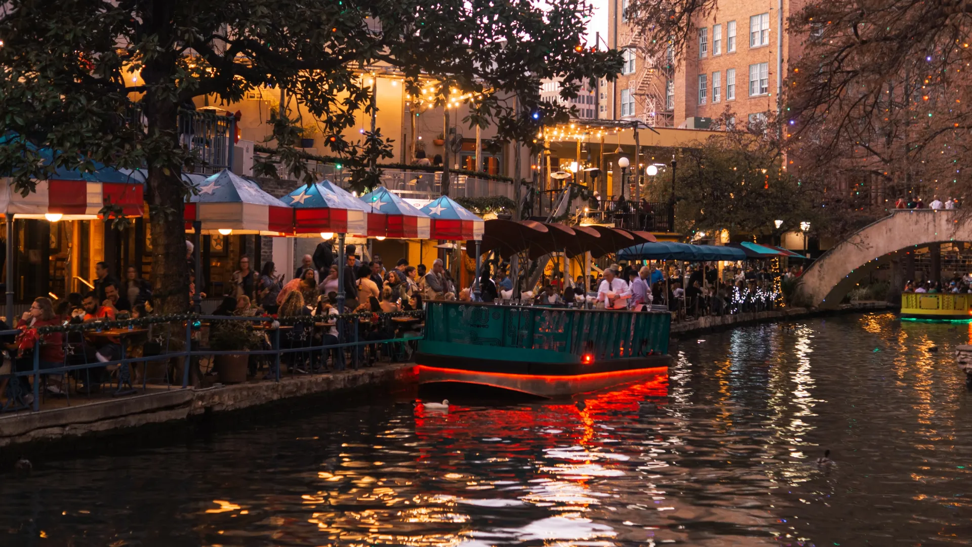 a boat full of people on a river with San Antonio River Walk in the background