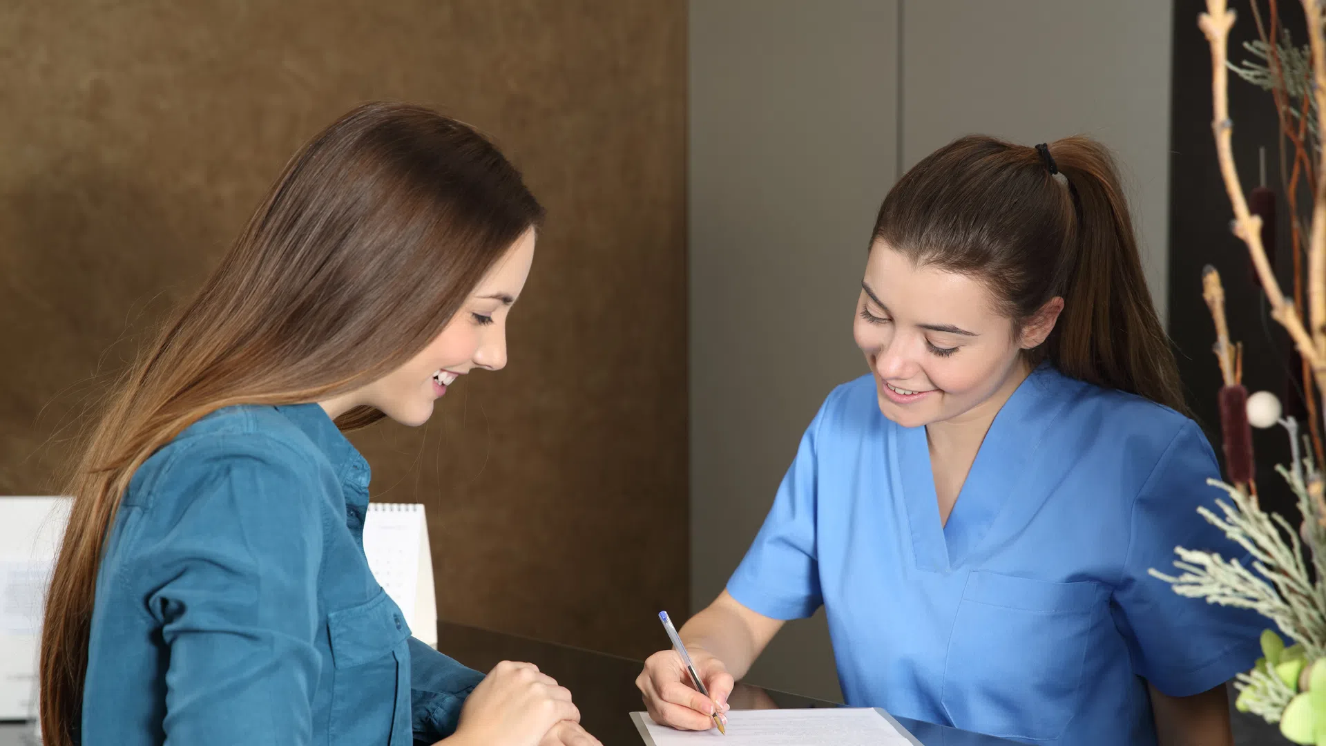 Healthcare professional in blue scrubs writing on clipboard while patient smiles during consultation