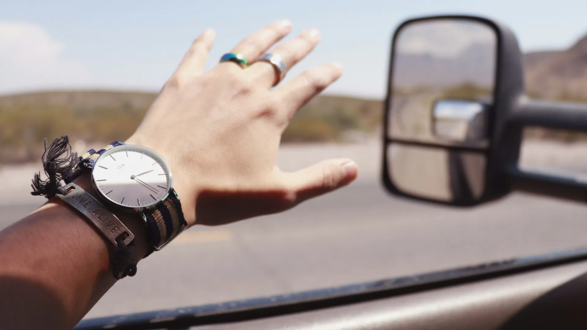 Person's hand with watch and rings extended out of car window with side mirror and desert landscape.