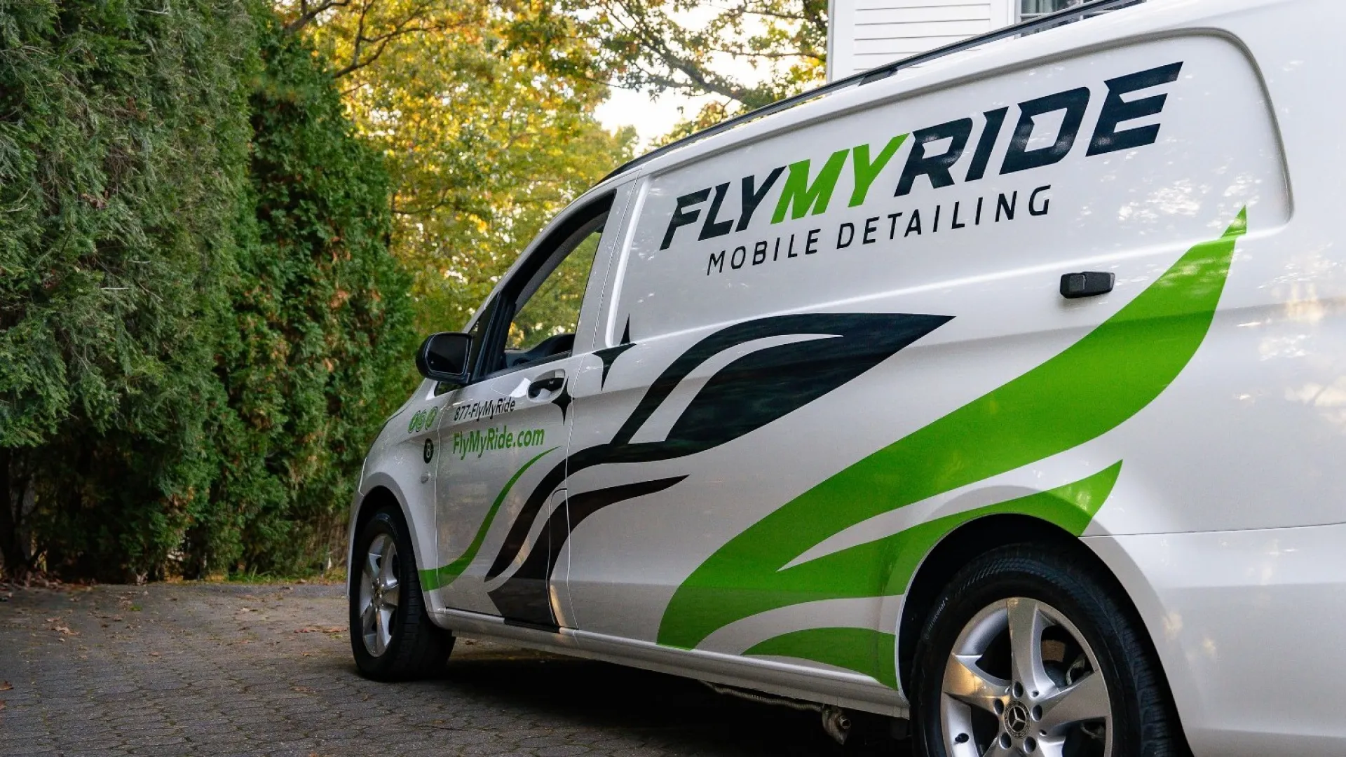White Fly My Ride mobile detailing van with green and black graphics parked on driveway near trees