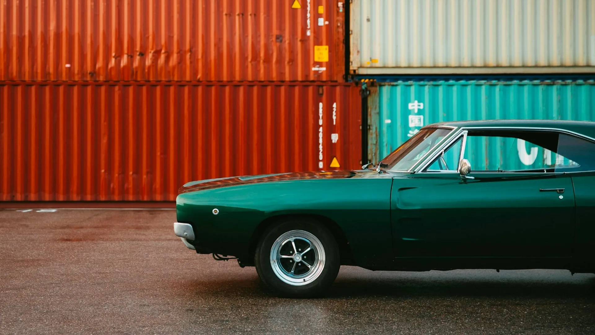 Green vintage car parked in front of stacked rust-colored and light blue shipping containers