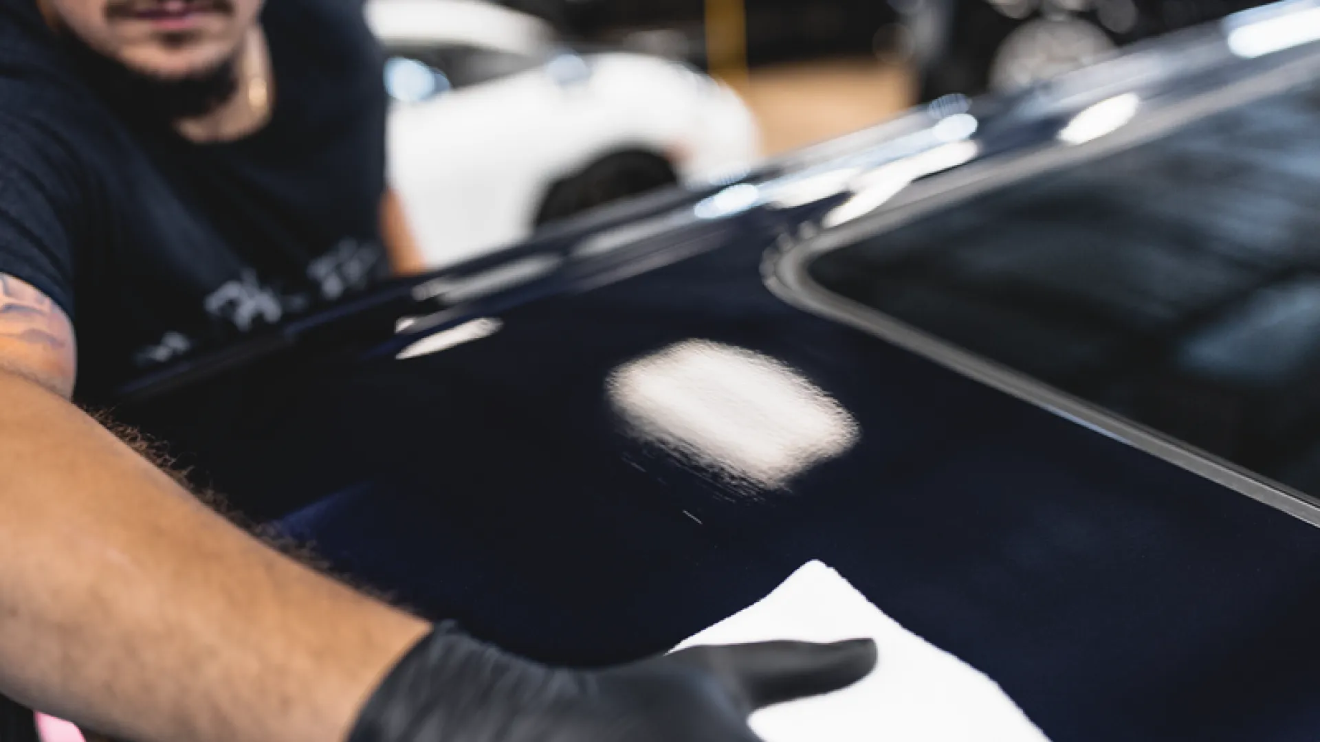 Person wearing black gloves polishing a shiny black car hood with a white cloth in a garage.