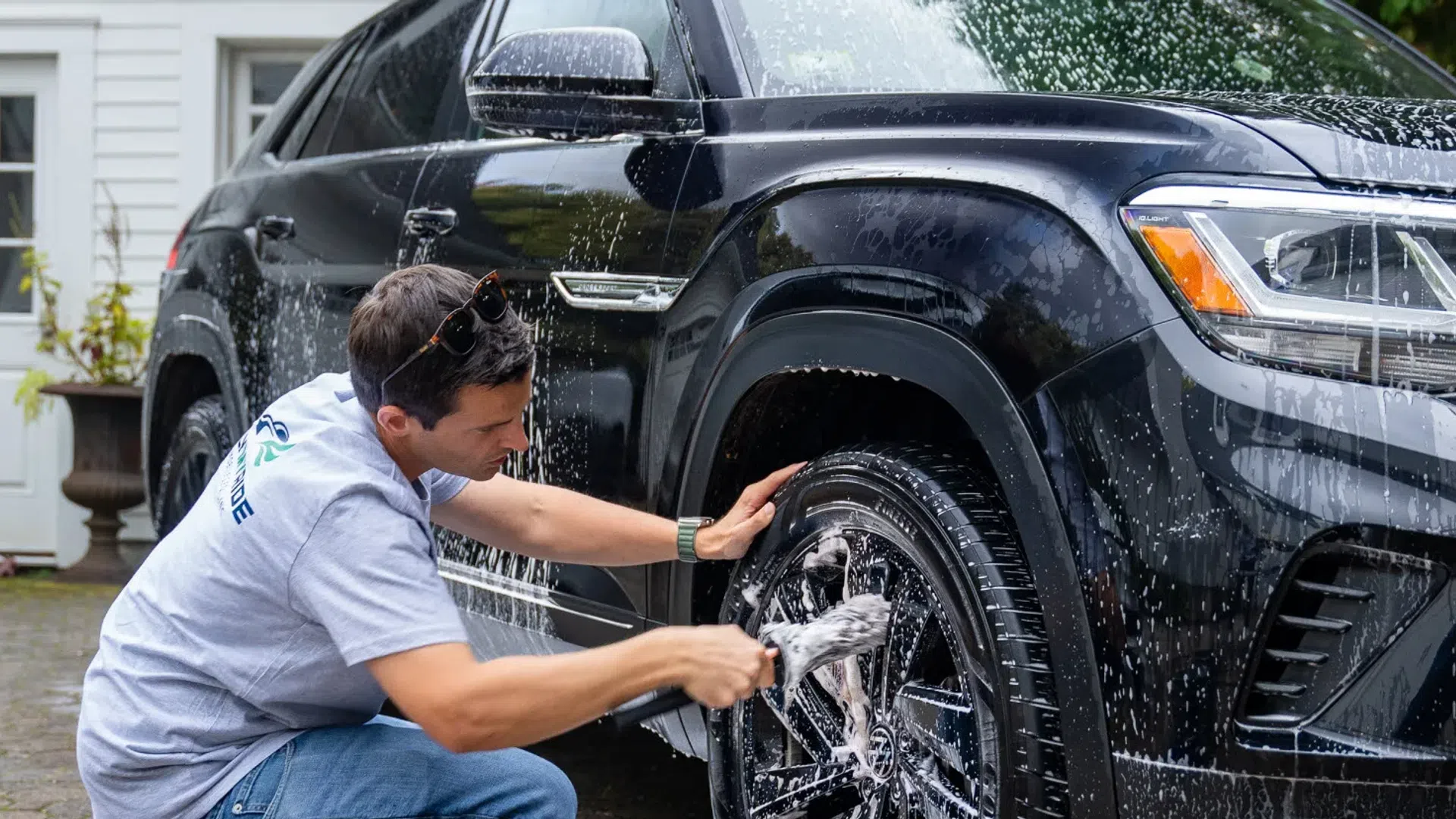 Man washing the tire and rim of a black SUV with soap and water in a driveway outdoors.