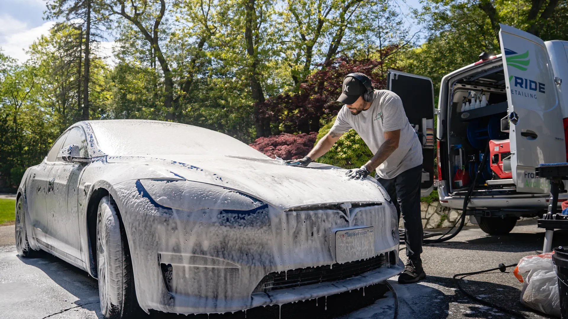 Man washing a Tesla car covered in foam beside a detailing van on a sunny day.