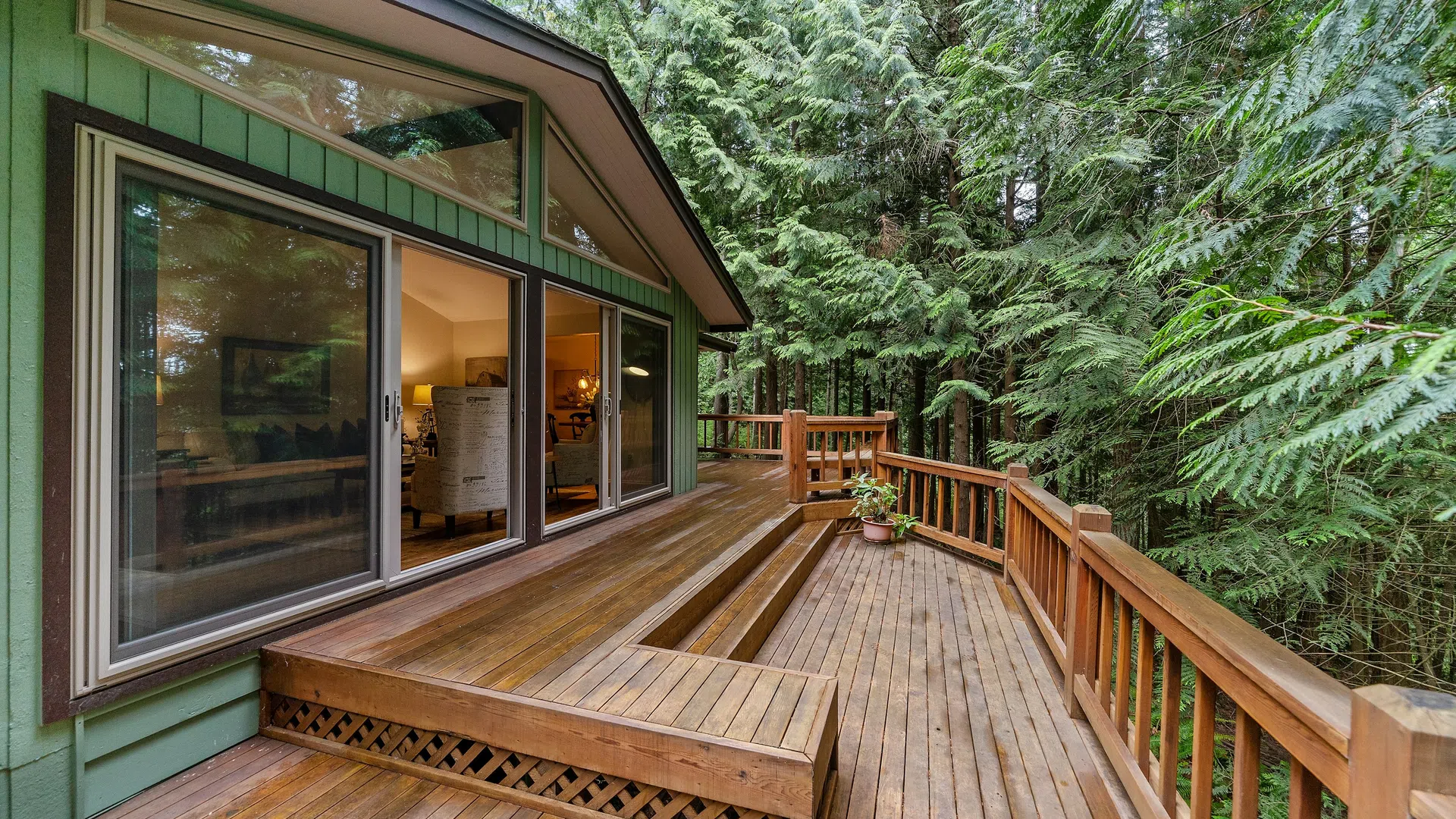 Wooden deck with built-in seating attached to green house surrounded by tall trees and plants.
