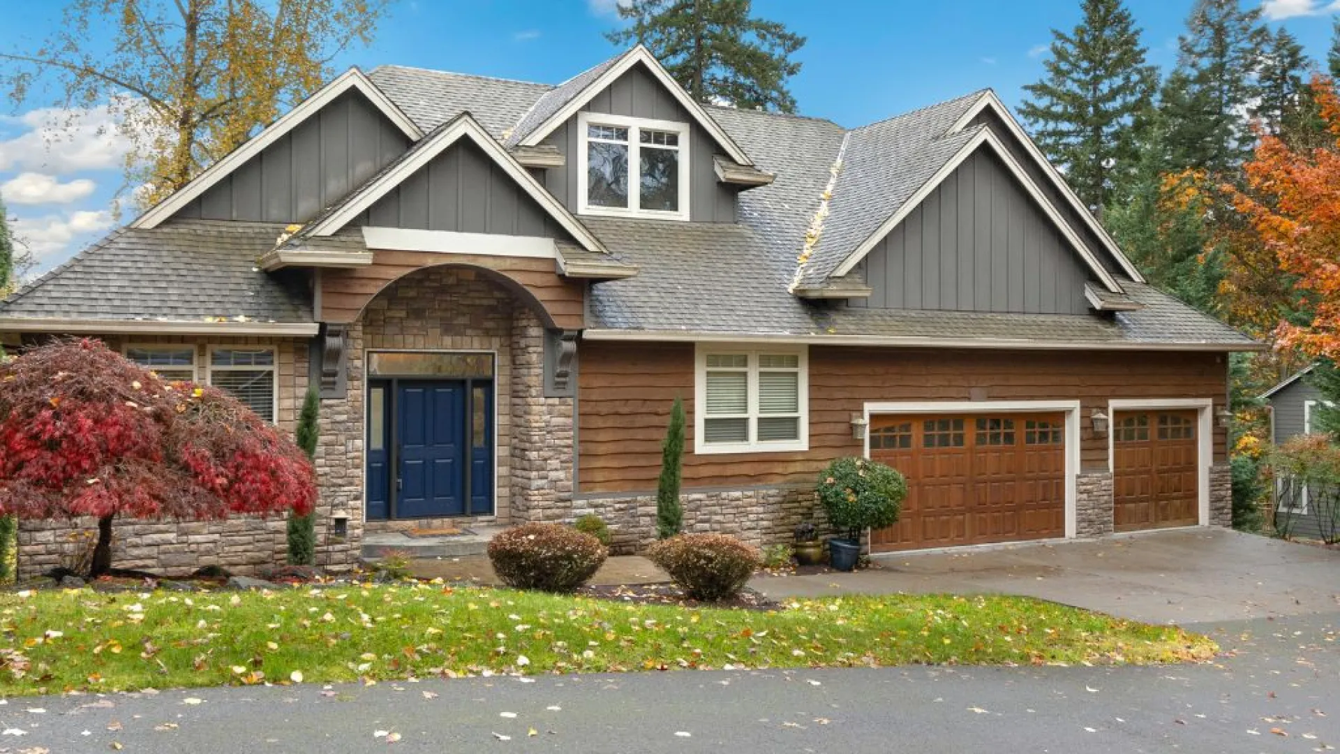Modern house exterior with gray roof, stone and wood siding, blue front door, three-car garage, and landscaped yard.
