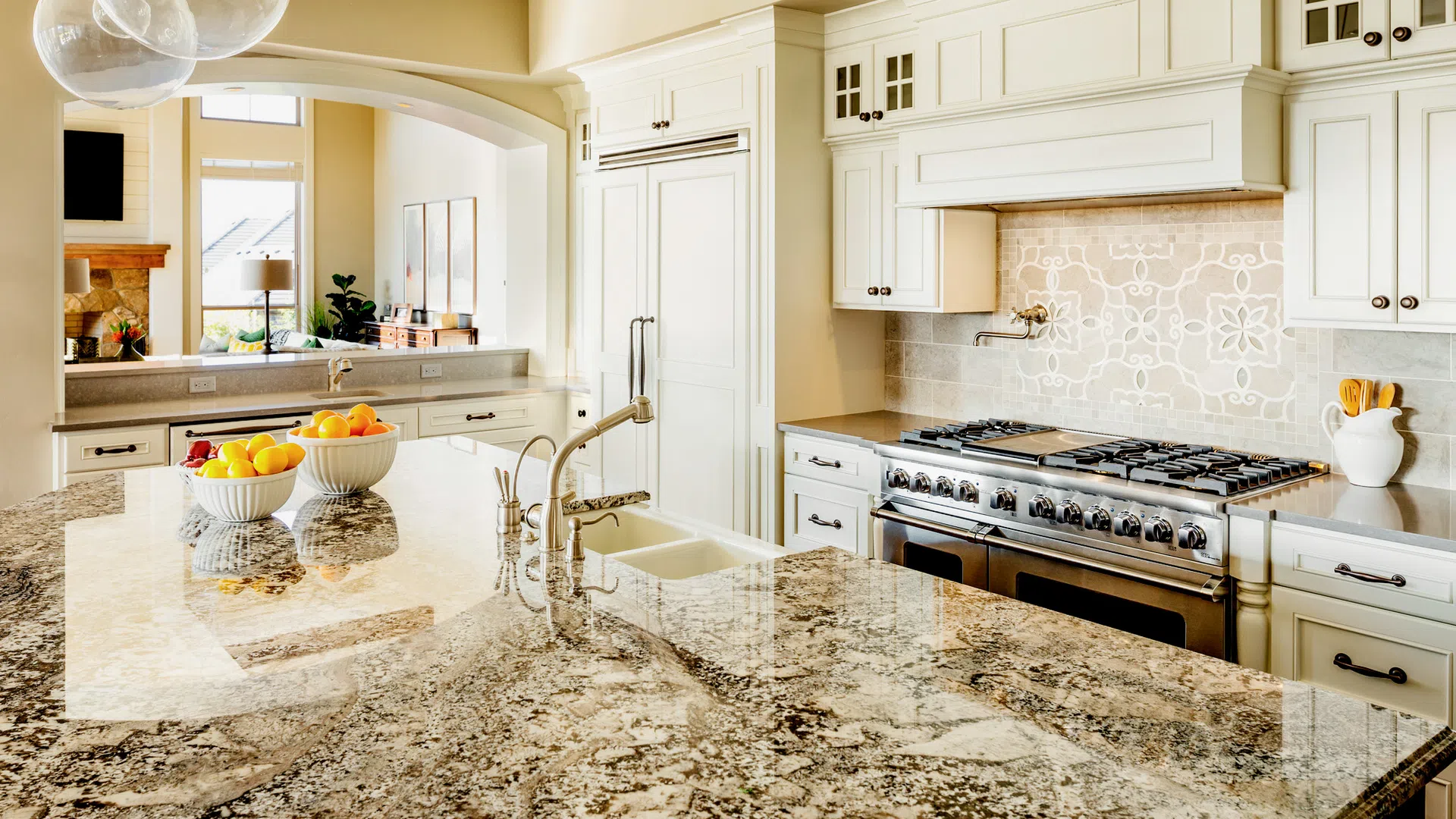 Bright modern kitchen with granite island, white cabinets, stainless steel stove, and bowls of fruit near window.