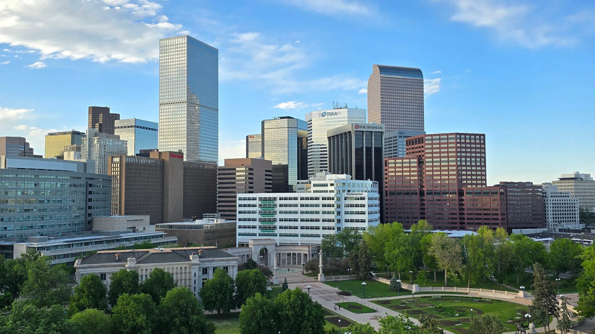 Downtown city skyline with modern skyscrapers and a green park under a partly cloudy blue sky