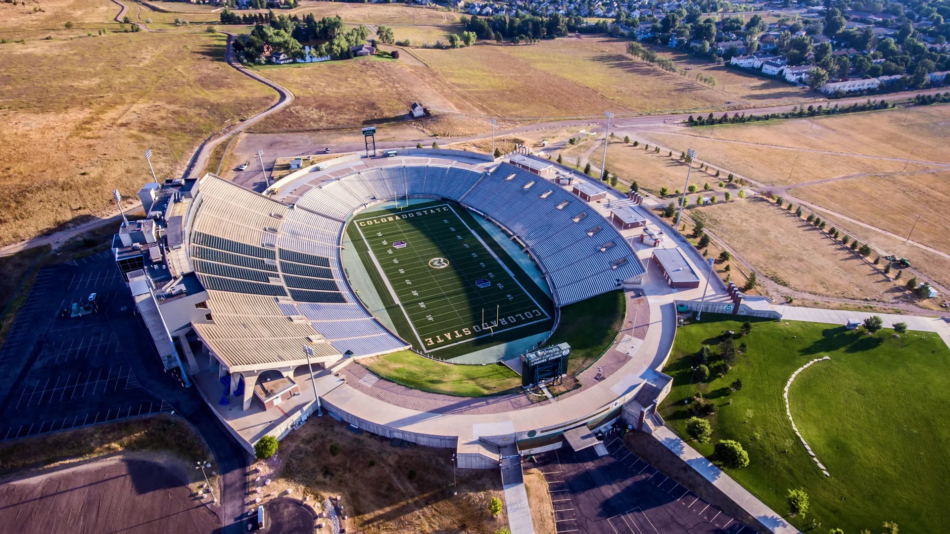 Aerial view of an empty Colorado State University football stadium surrounded by dry fields and nearby residential area.