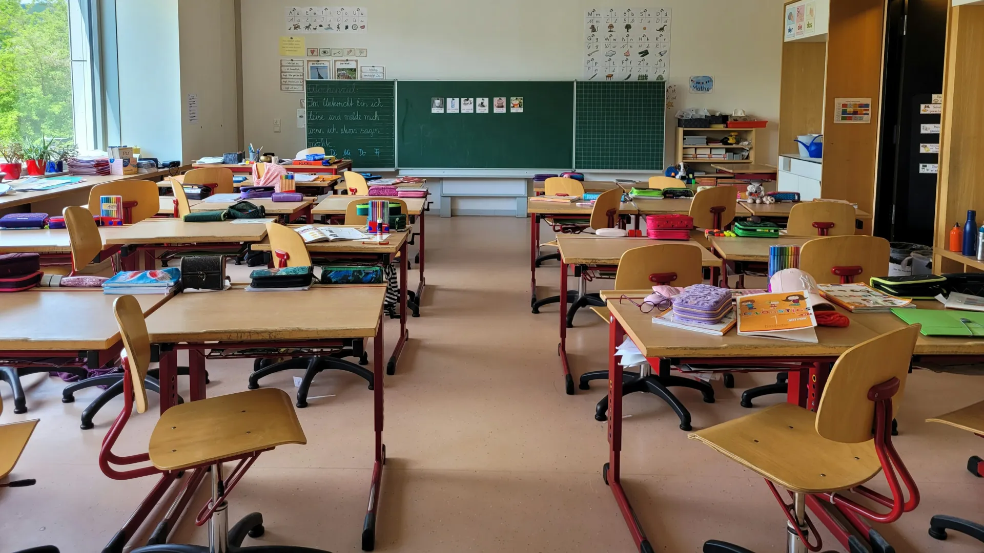 Bright, organized classroom with wooden desks, chairs, books, and a large chalkboard at the front.