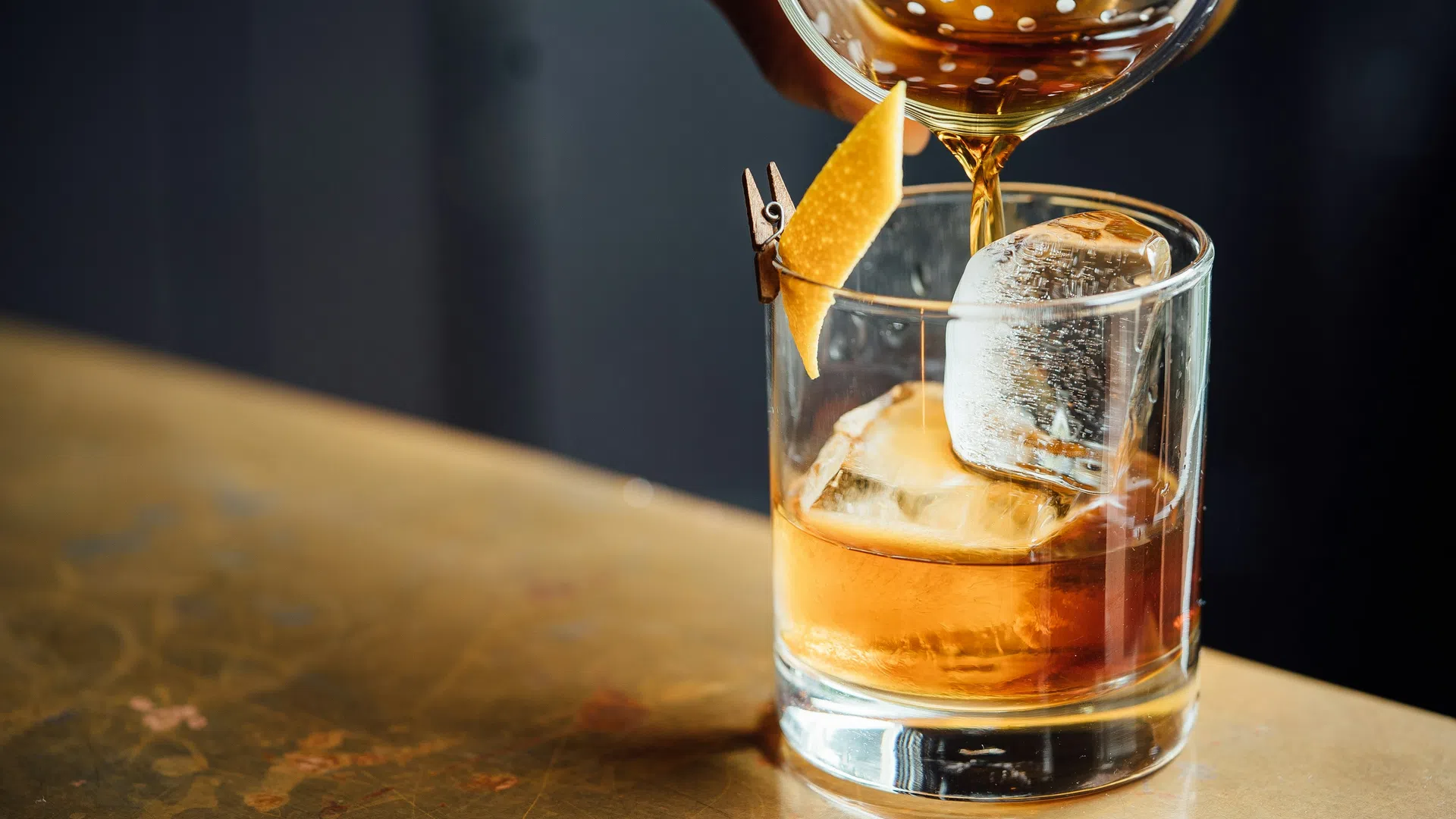 Close-up of a cocktail being poured over ice with a lemon twist garnish on a polished surface.