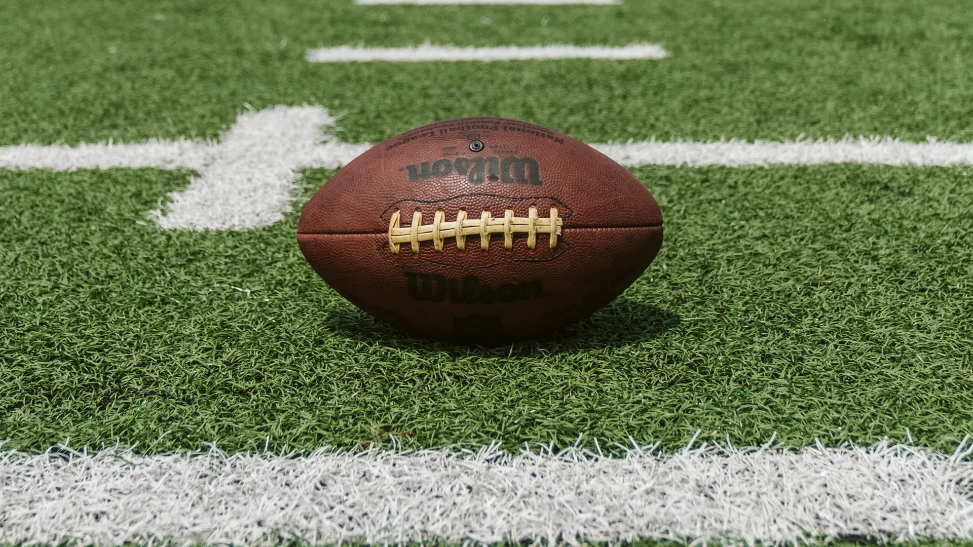 Wilson football placed on green turf field near white yard lines ready for play.