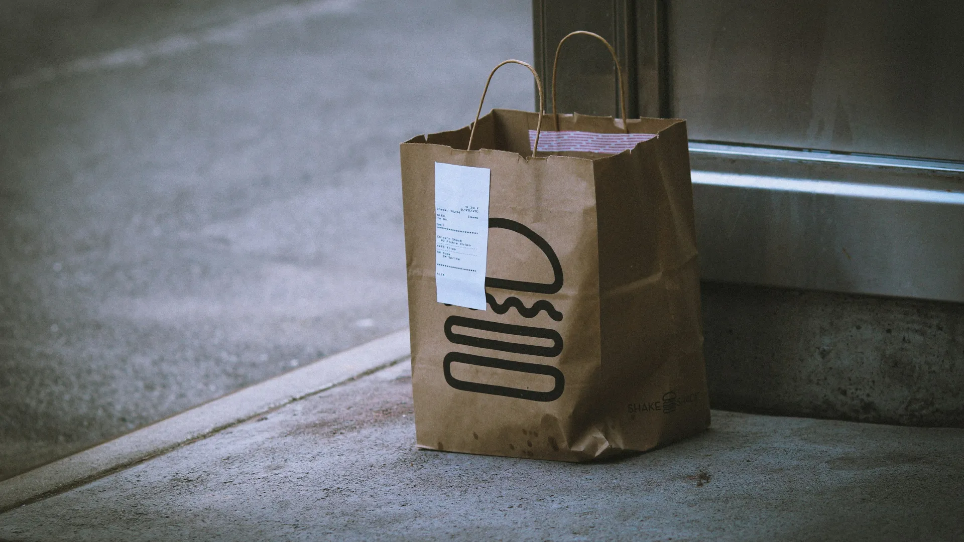 Brown paper bag with a burger logo and receipt left on a concrete sidewalk, likely a food delivery or takeout.