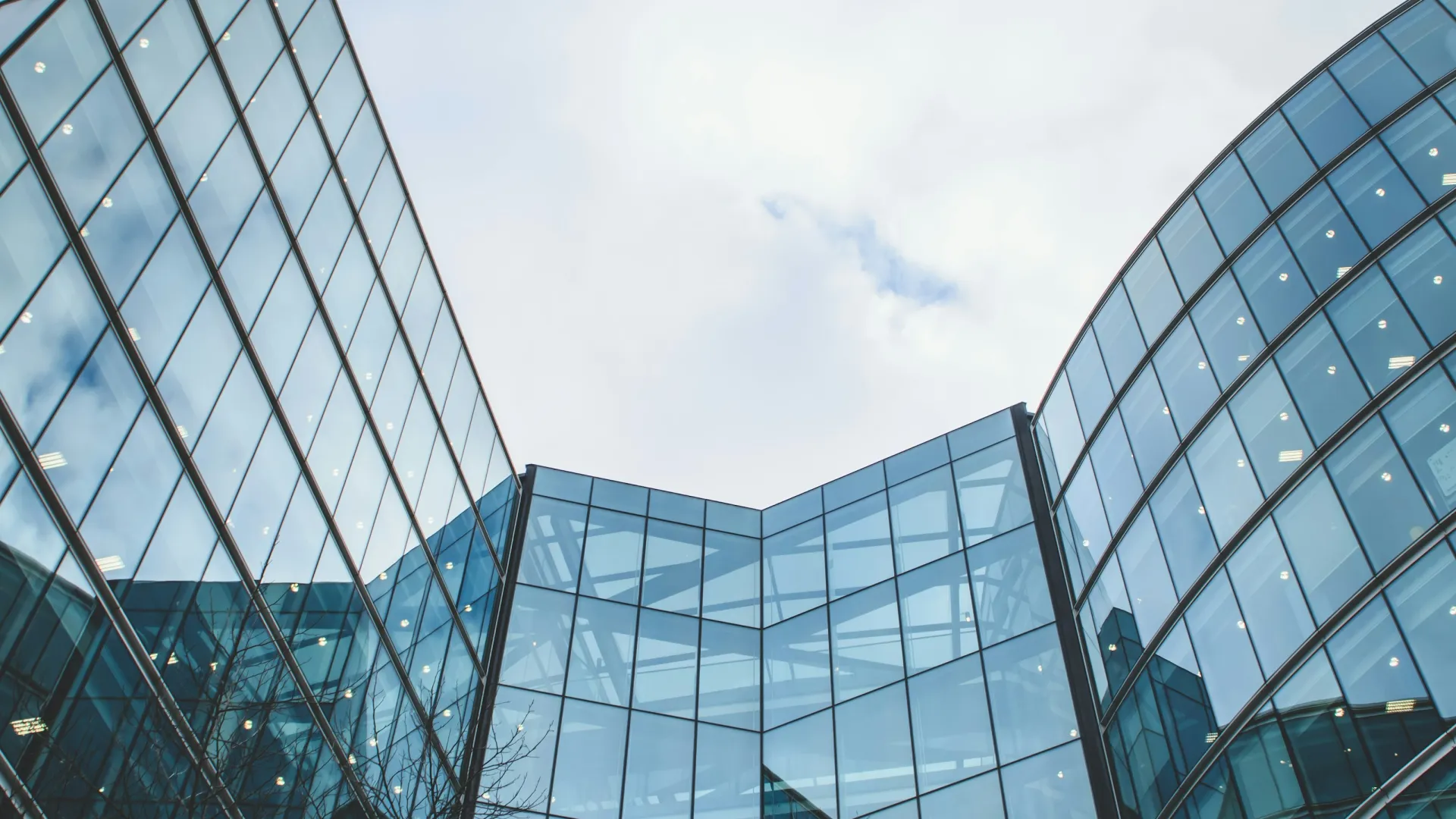 Modern glass office buildings with reflections of sky and leafless trees on a cloudy day.