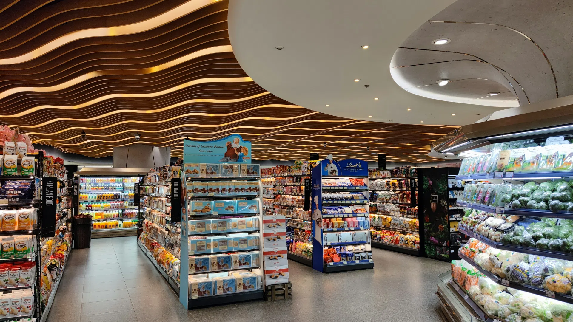 Modern grocery store aisle with wavy wooden ceiling, refrigerated produce, and stocked shelves under bright lights