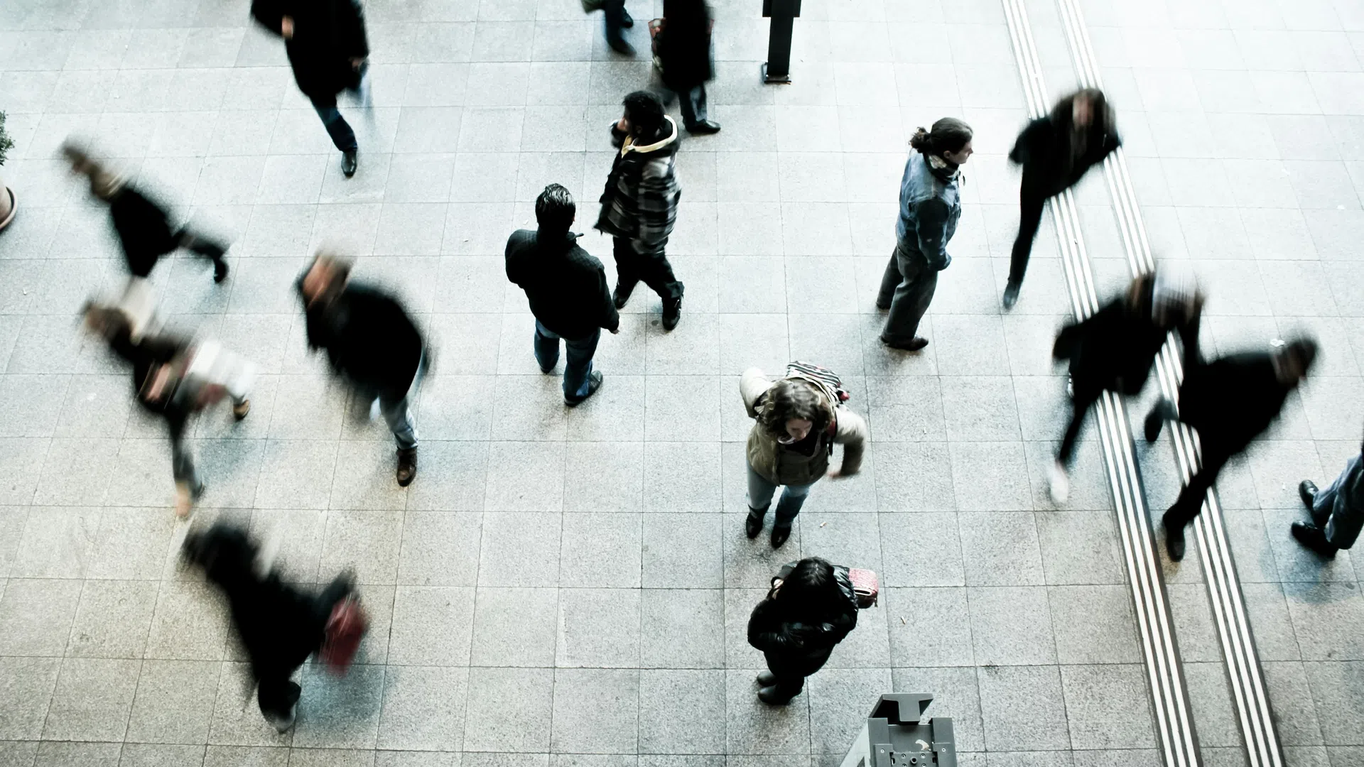 Overhead view of people walking and standing on tiled floor, some in motion blur, in urban public space.