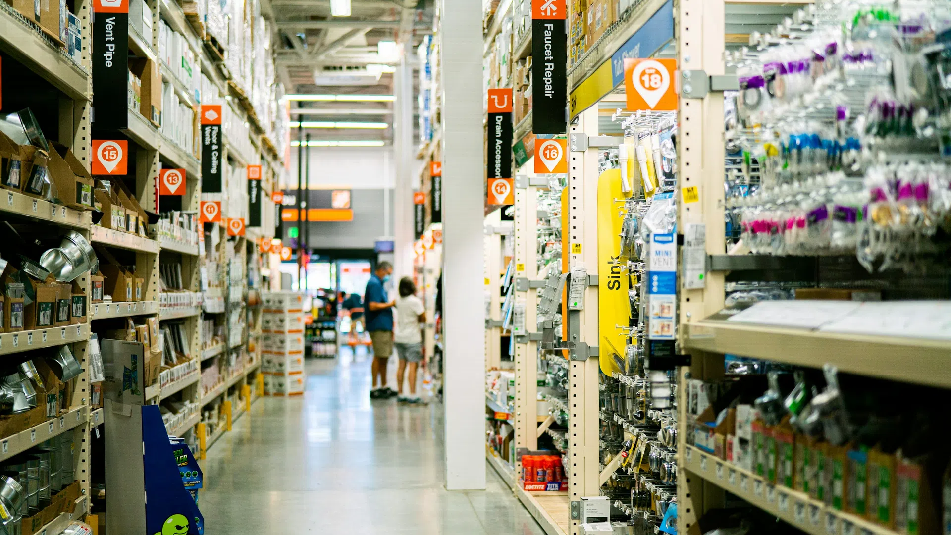 Aisle in a hardware store with shelves stocked with plumbing supplies and two shoppers in the background.