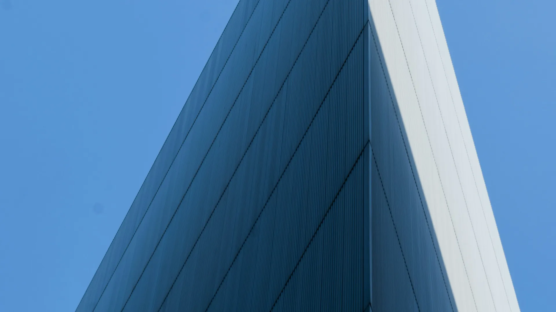 Sharp angled modern building facade against clear blue sky with contrasting light and shadows.