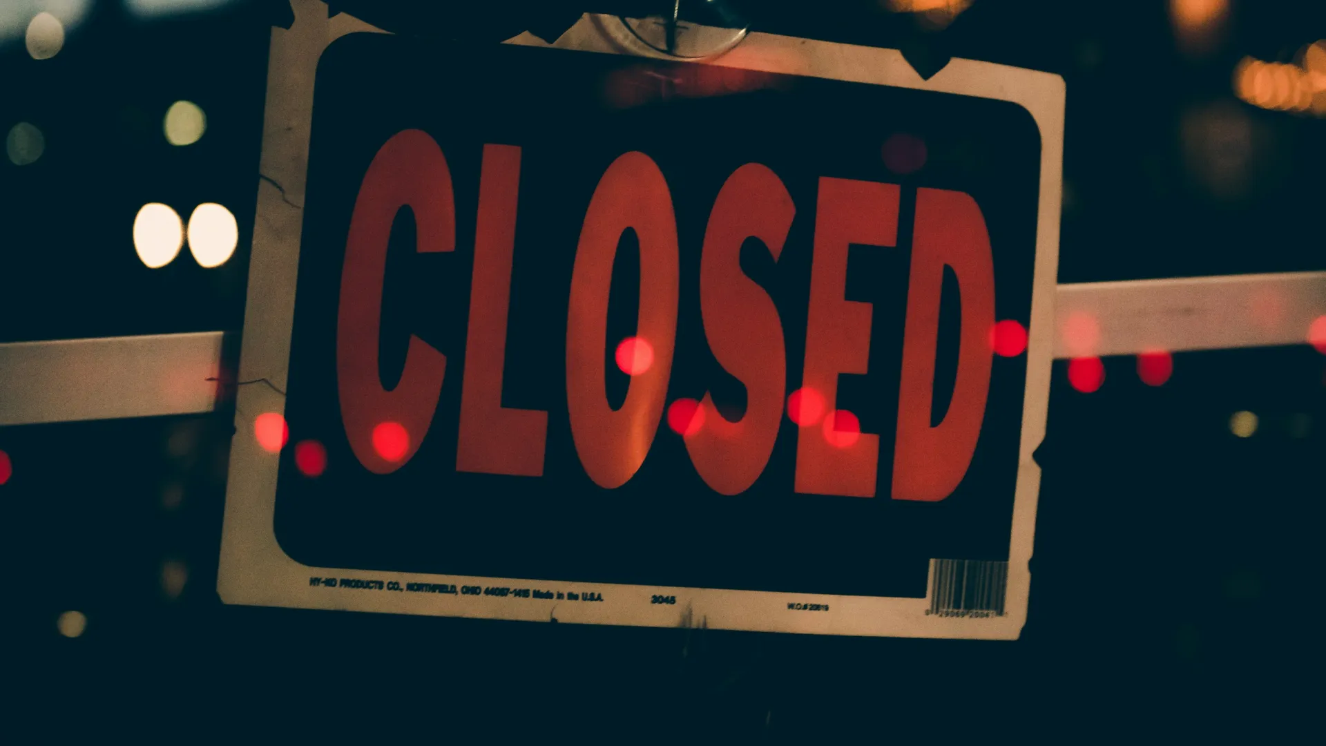 Red closed sign hanging on a door with blurred city lights in the dark background.