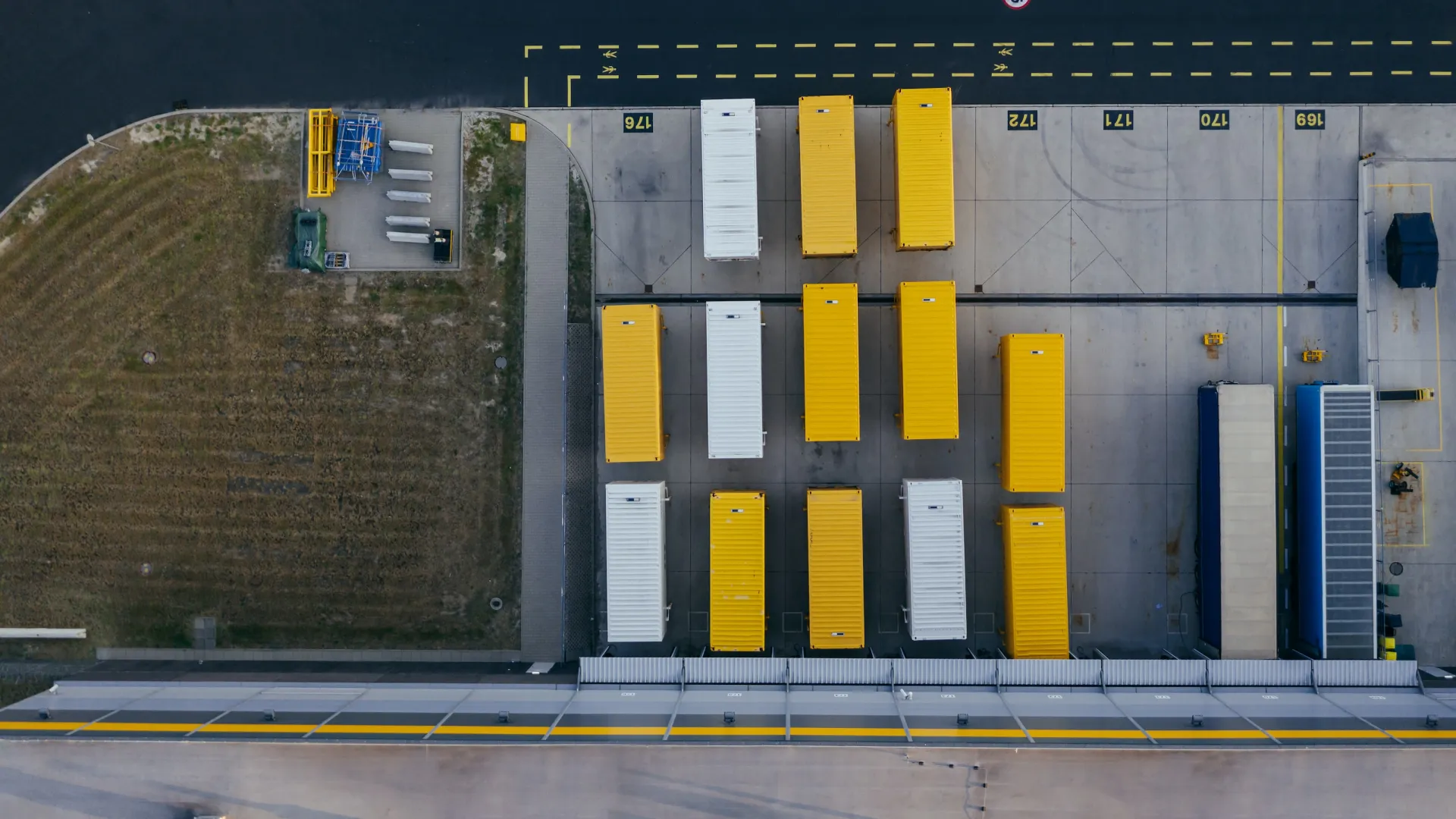 Aerial view of shipping containers, mostly yellow and white, neatly arranged in a concrete storage yard next to a grassy area.