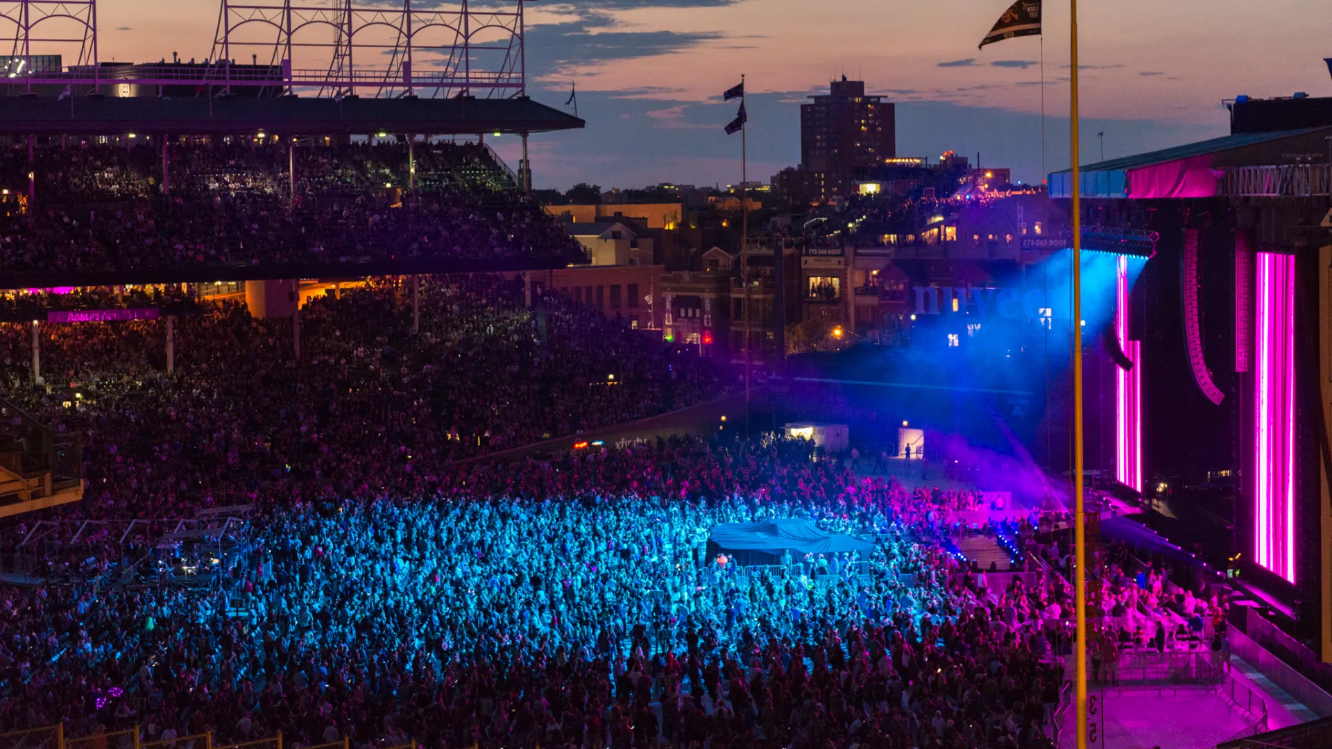 Large crowd at an outdoor concert at dusk with colorful stage lights and city skyline in the background.