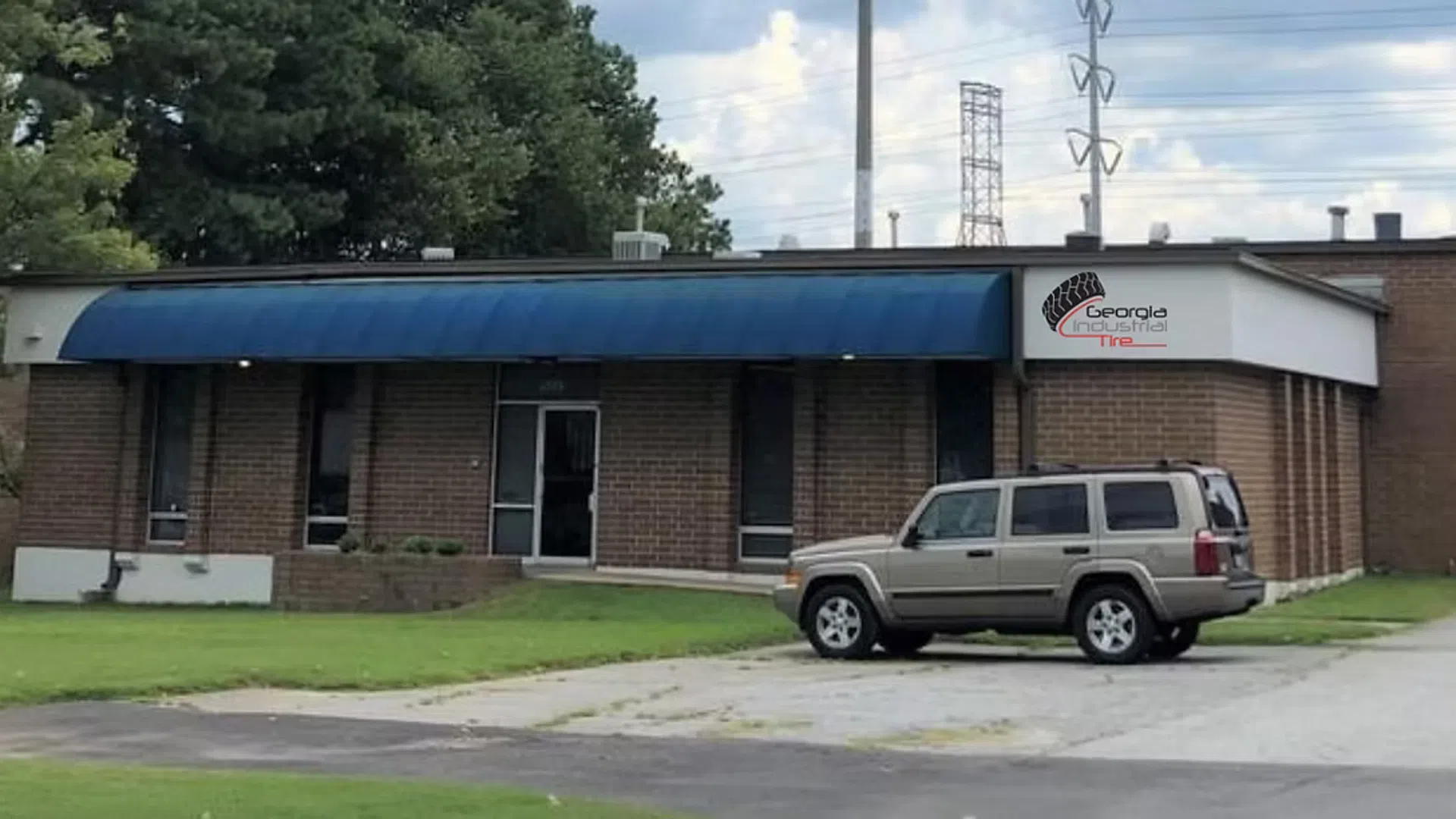 Brick office building with blue awning, vehicles parked outside, and grassy lawn under cloudy sky.