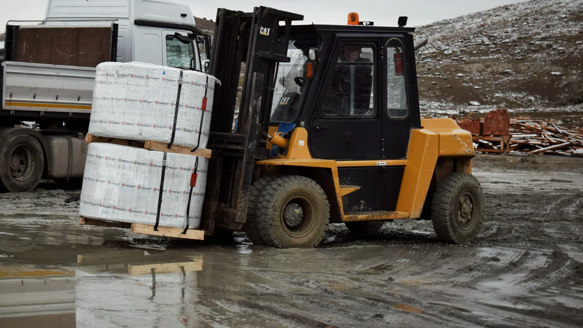 Yellow forklift lifting wrapped pallets in an outdoor muddy industrial area with a white truck in the background.