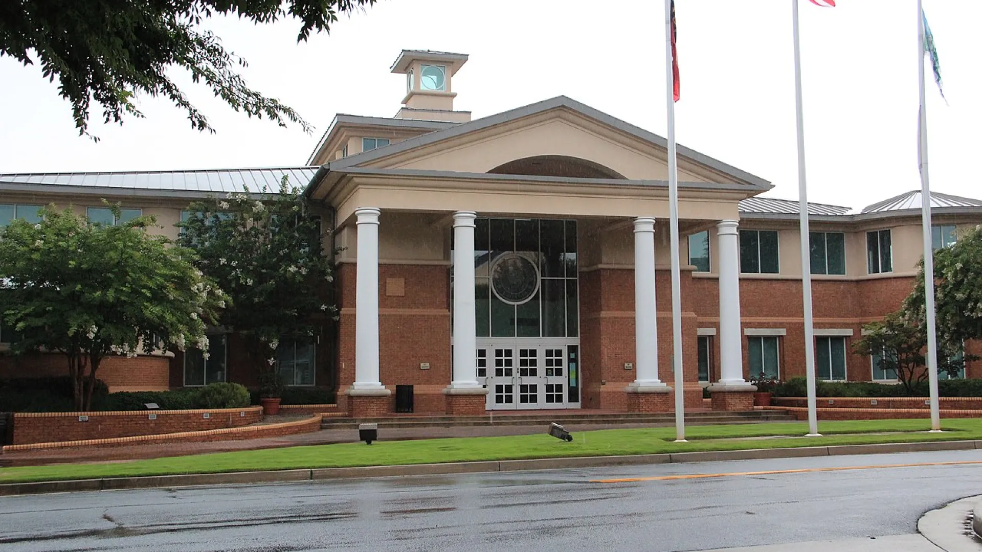Front view of a government or municipal brick building with white columns and flags on a rainy day