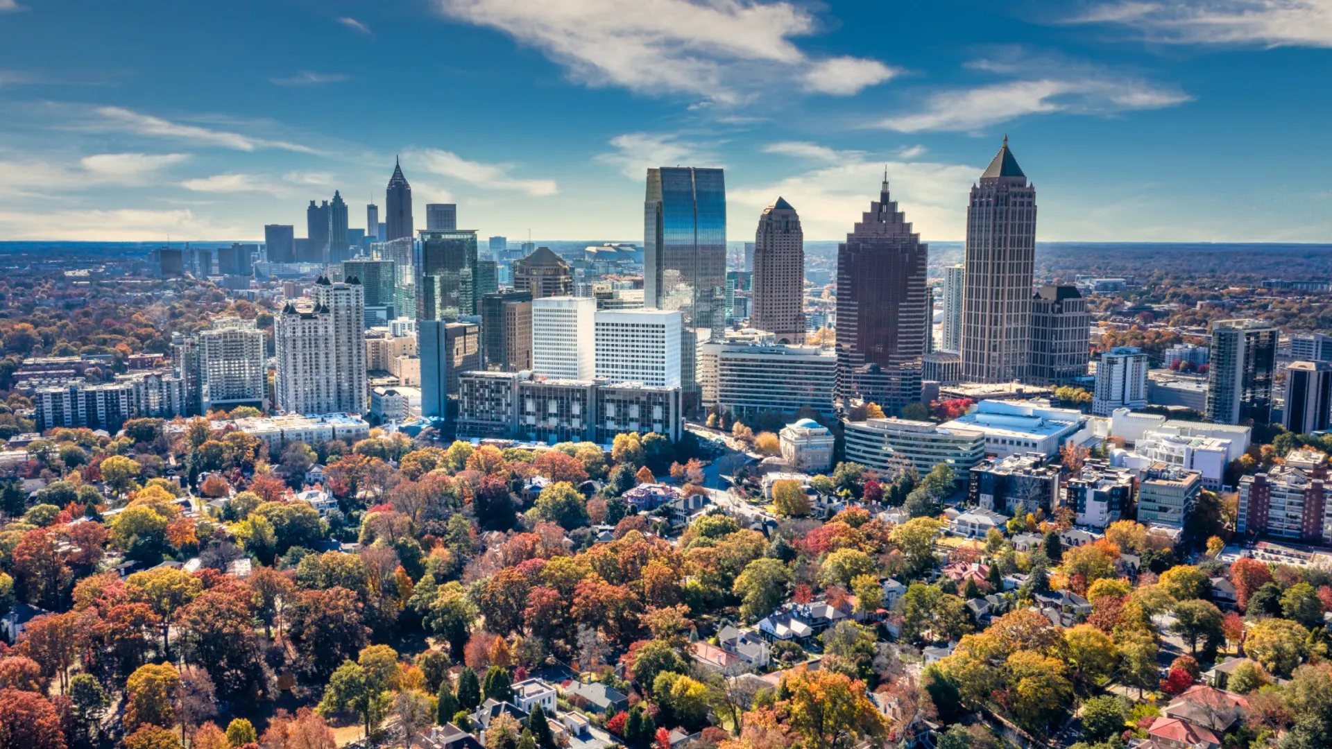 Aerial view of a city skyline with skyscrapers and colorful autumn foliage in the foreground under a blue sky.